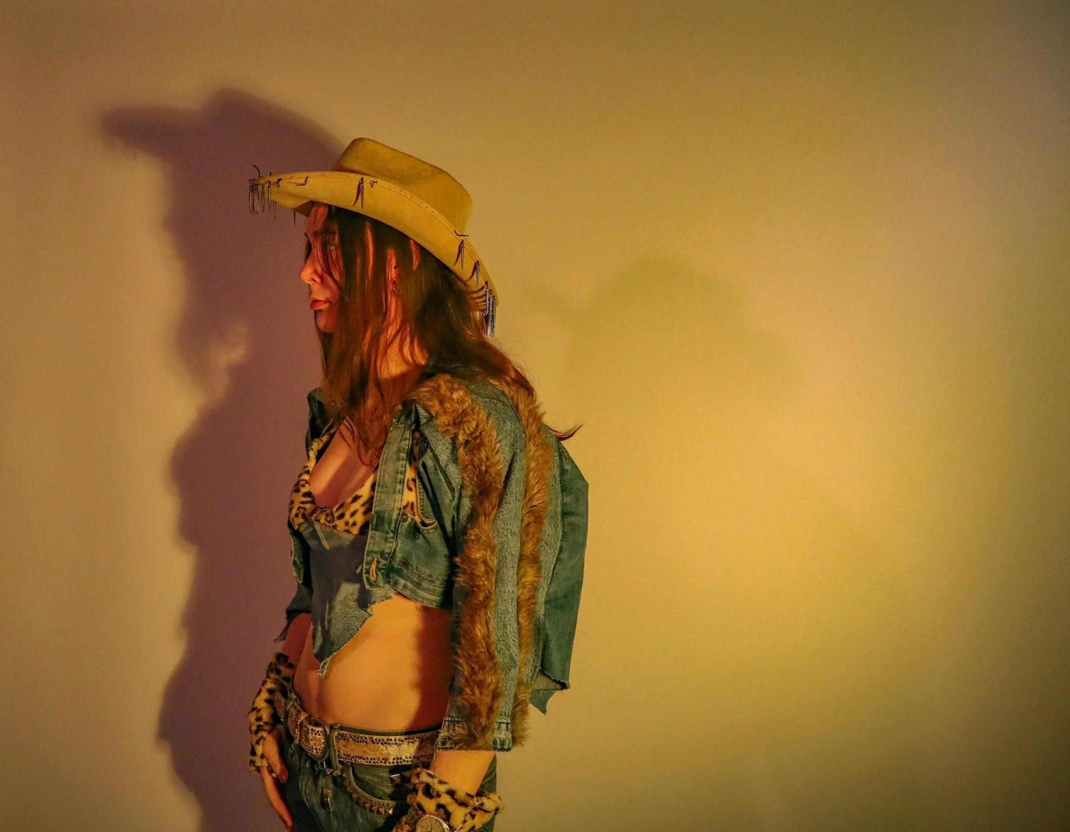A woman with long hair wearing a yellow cowboy hat adorned with metal embellishments, a leopard-print top, and a denim jacket with fur trim, standing sideways against a plain yellow wall.
