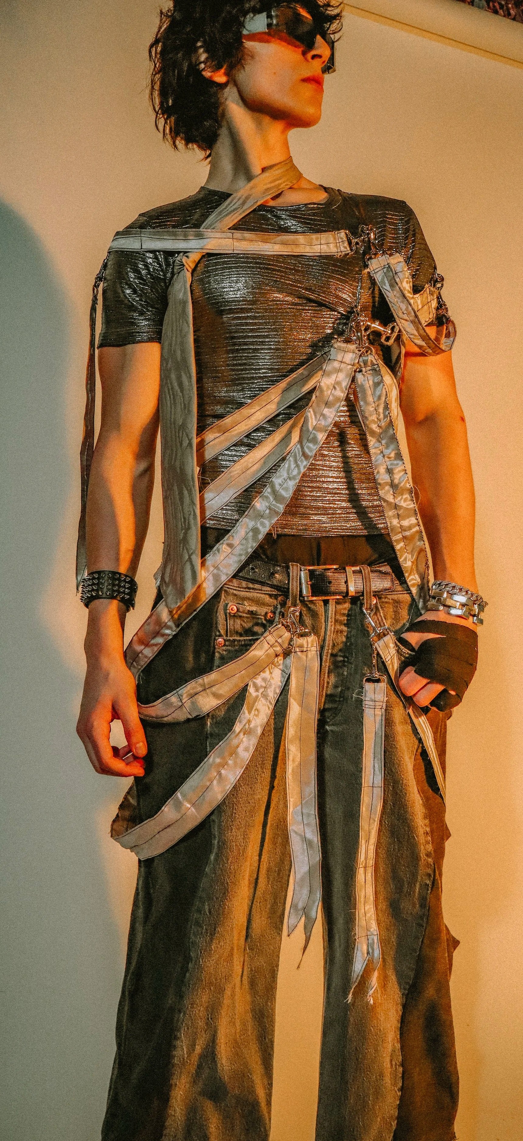 A person with curly dark hair wearing large futuristic silver reflective sunglasses, a metallic silver textured shirt, and a layered outfit with straps, chains, and accessories, standing against a plain background with yellow and blue lighting.