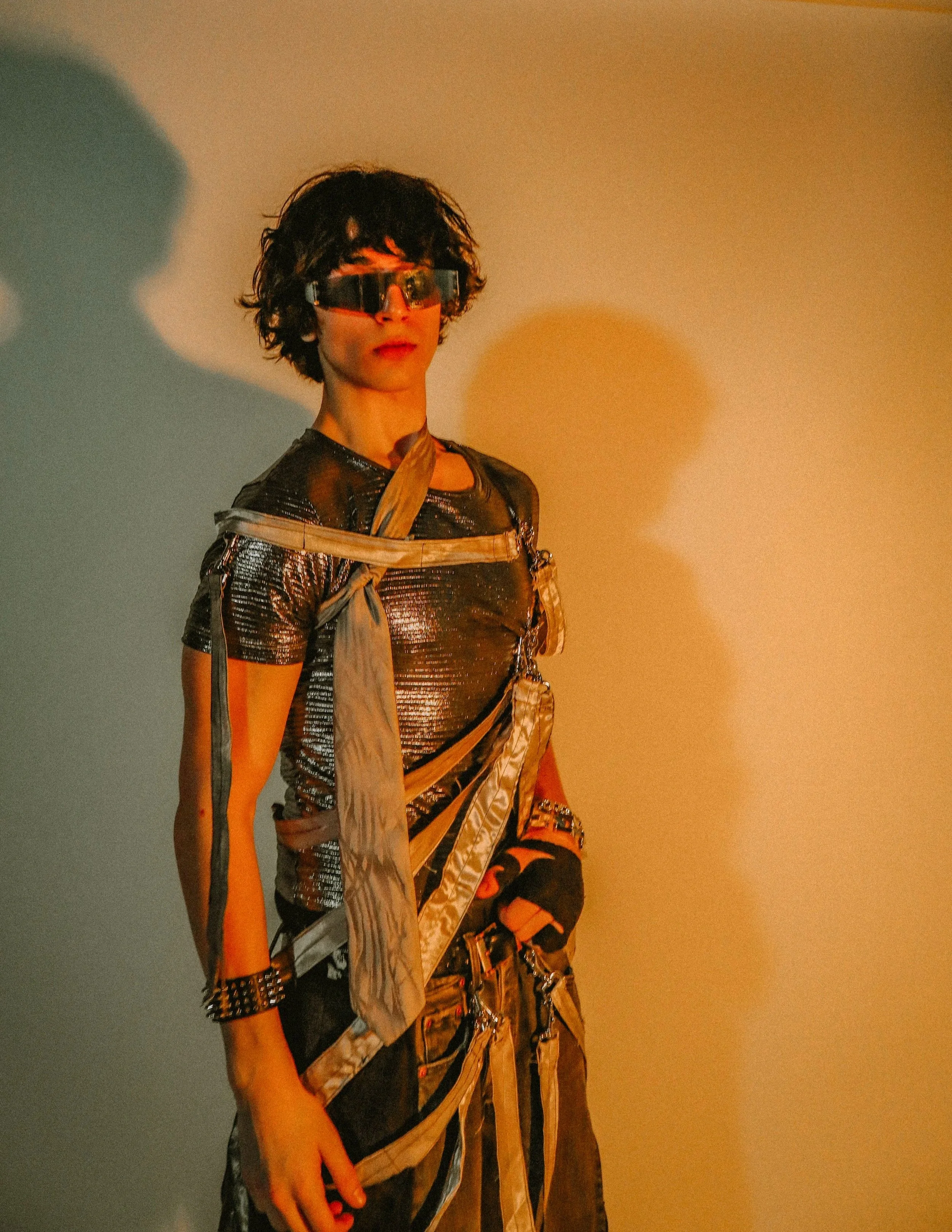 A person with short, curly dark hair wearing reflective sunglasses, a metallic shiny top, layered clothing, and studded accessories, standing against a multicolored background with shadows.