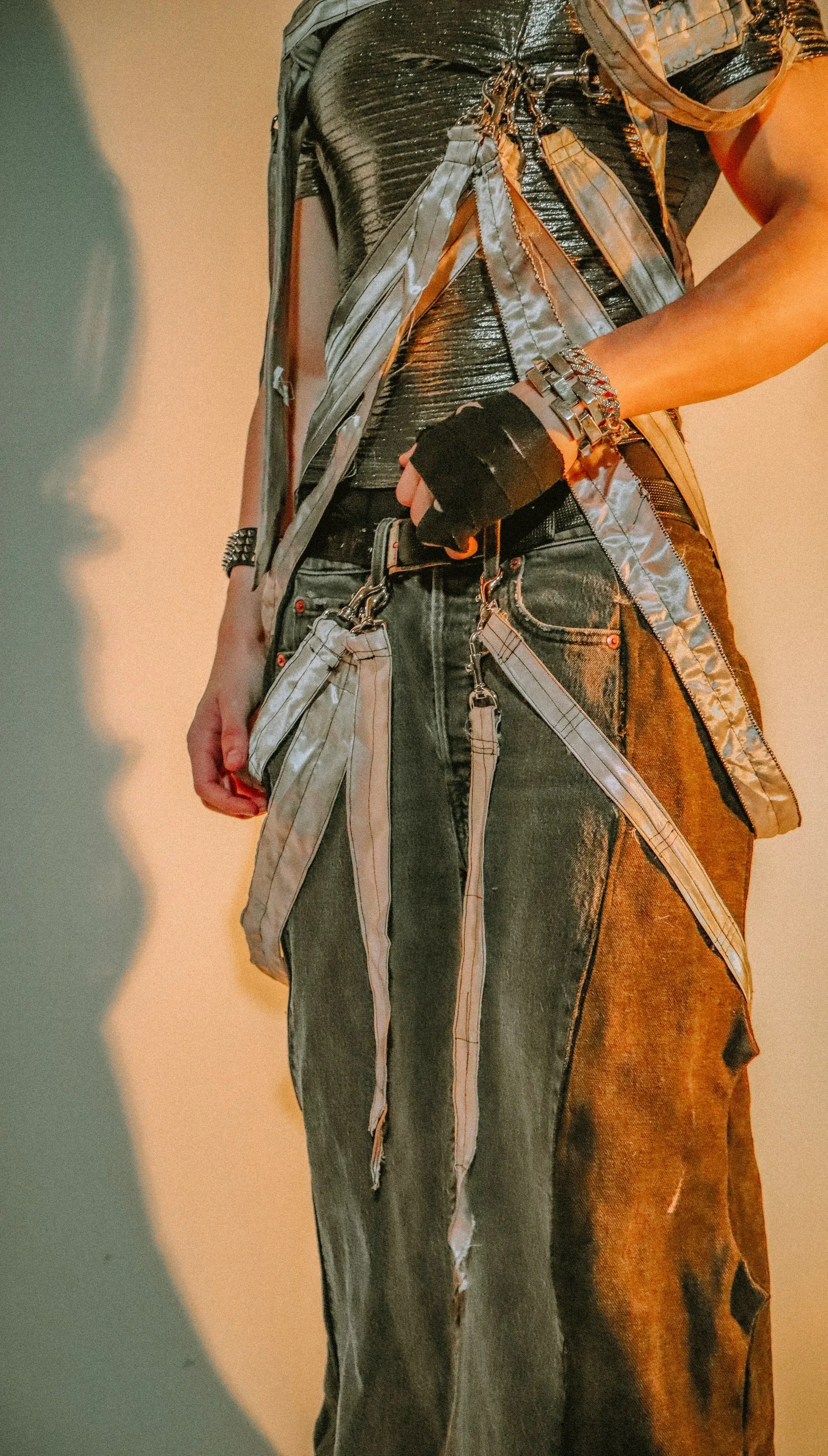 A person with curly dark hair wearing large futuristic silver reflective sunglasses, a metallic silver textured shirt, and a layered outfit with straps, chains, and accessories, standing against a plain background with yellow and blue lighting.