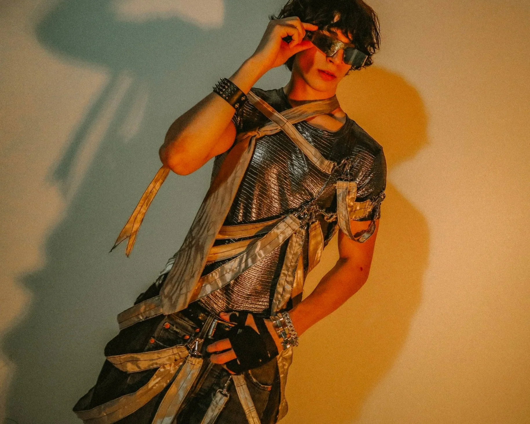 A person with curly dark hair wearing large futuristic silver reflective sunglasses, a metallic silver textured shirt, and a layered outfit with straps, chains, and accessories, standing against a plain background with yellow and blue lighting.