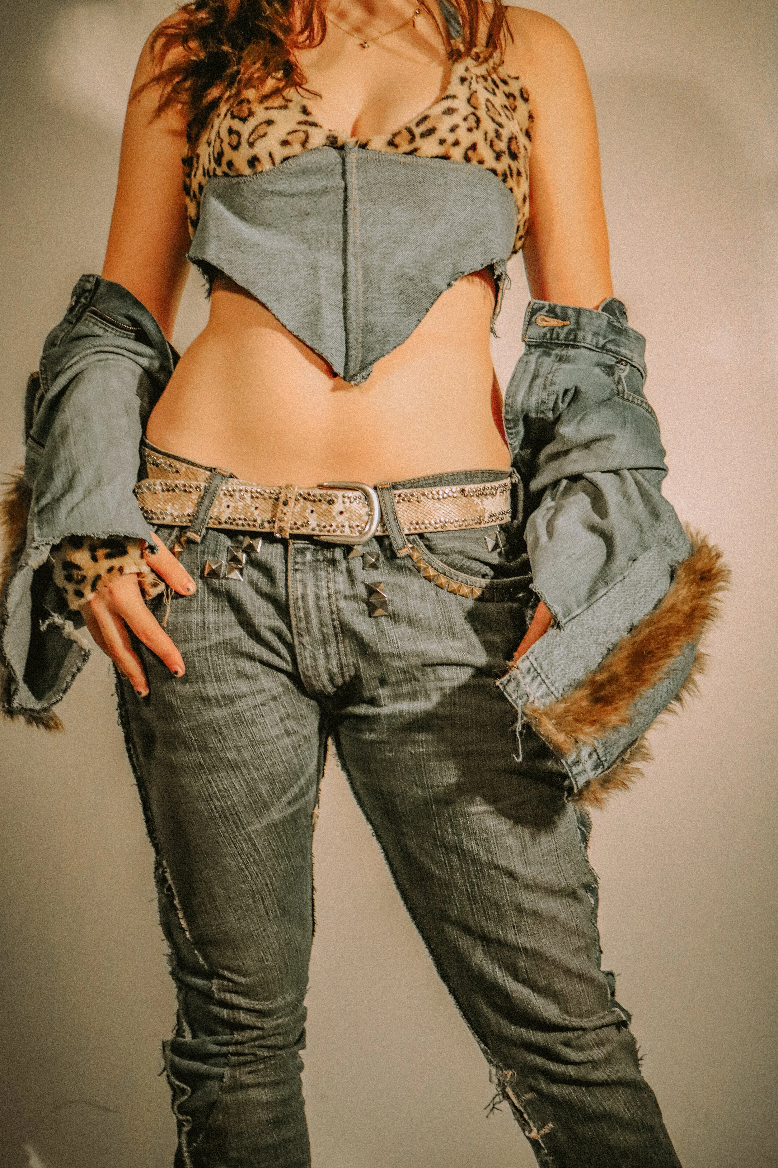 A person is wearing a leopard print top with black jeans, a studded belt, a denim jacket hanging off the shoulders, and a necklace. The person's face is not visible.
