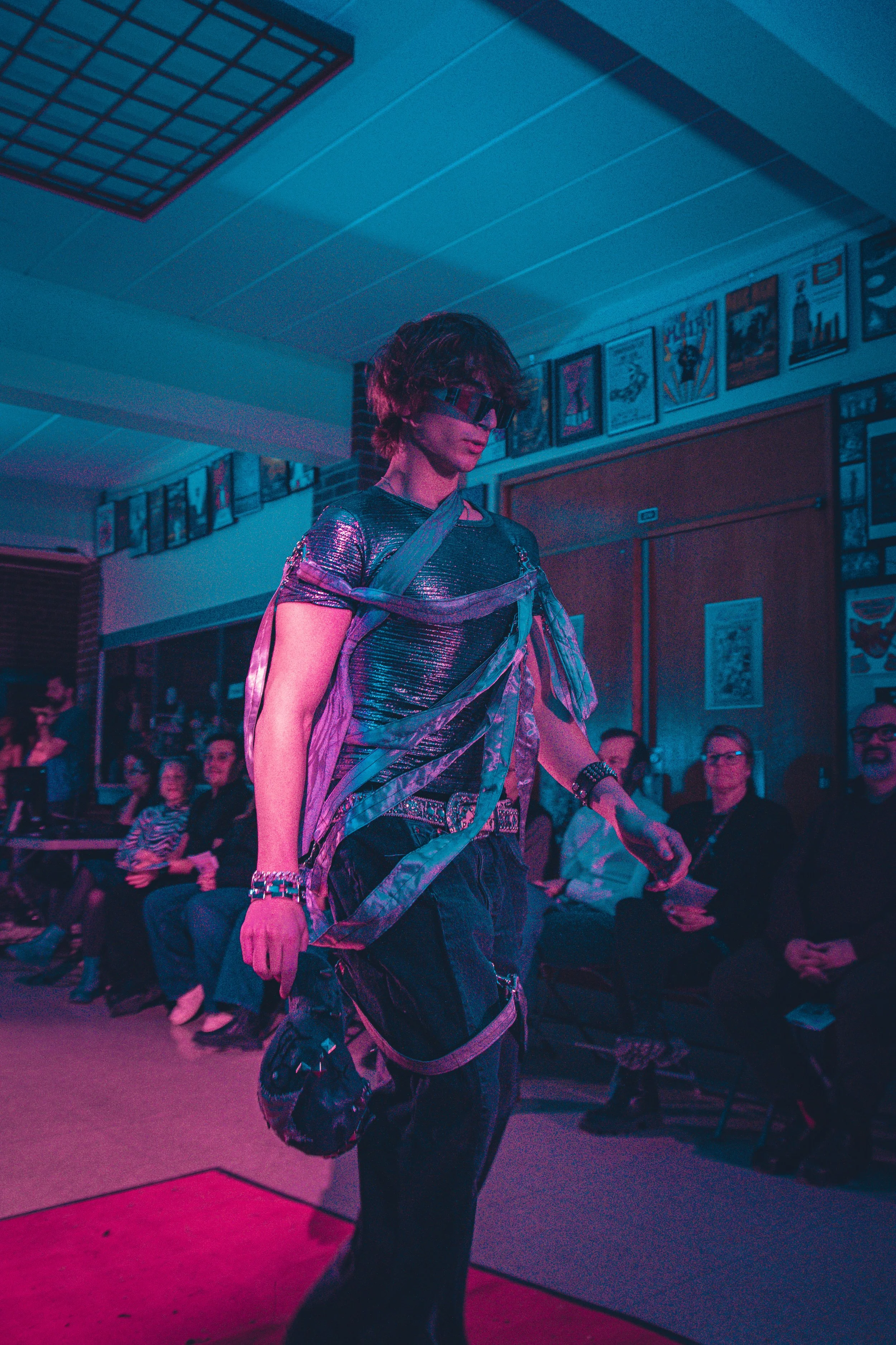 A person dressed in a shiny, metallic silver outfit with a matching hat, glasses, and gloves, walks on a runway in front of an audience, with blue lighting. Behind them are posters and a brick wall.
