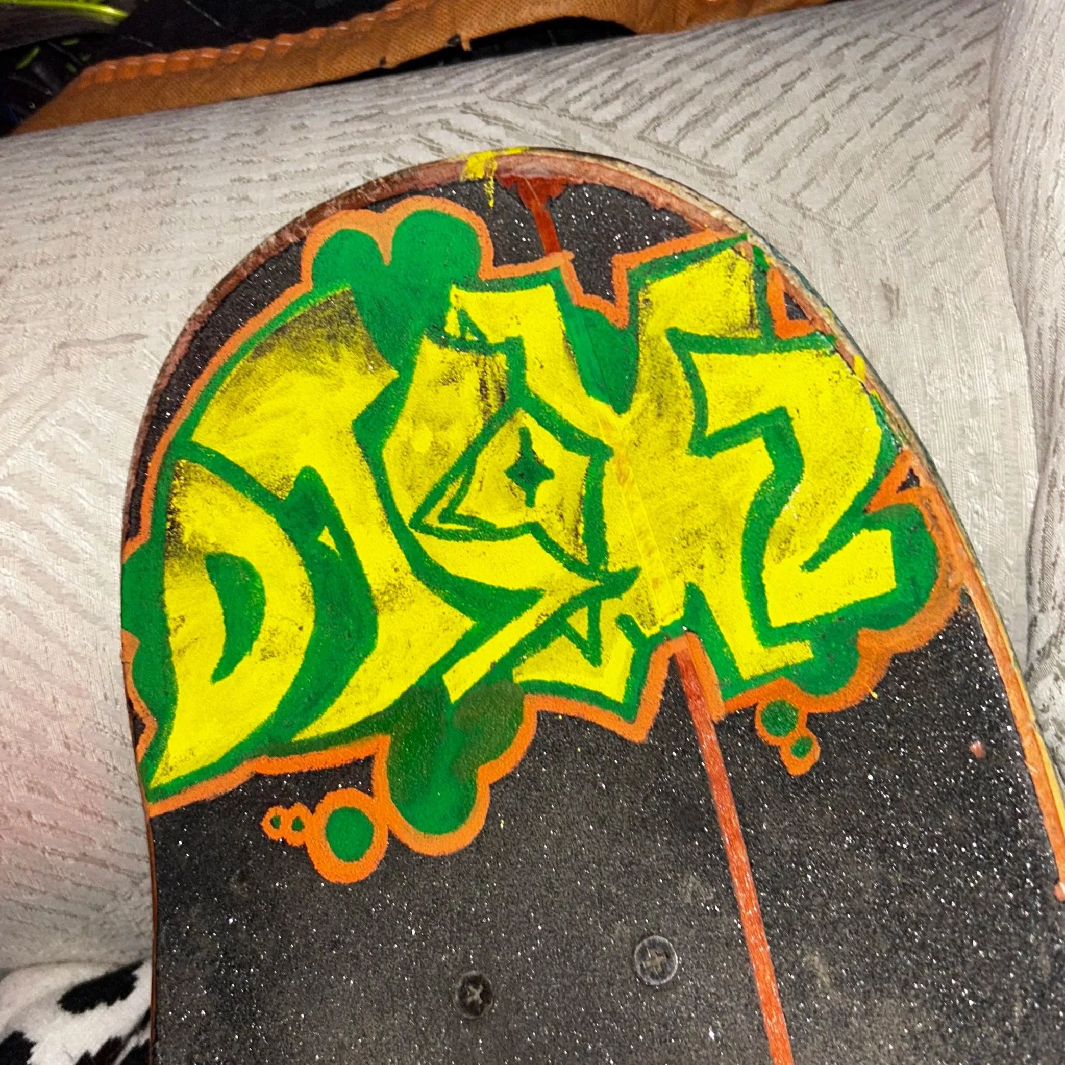 Part of a painted wooden surface with colorful graffiti-style art, including green, yellow, black, and orange elements, on a textured background with some visible screws.
