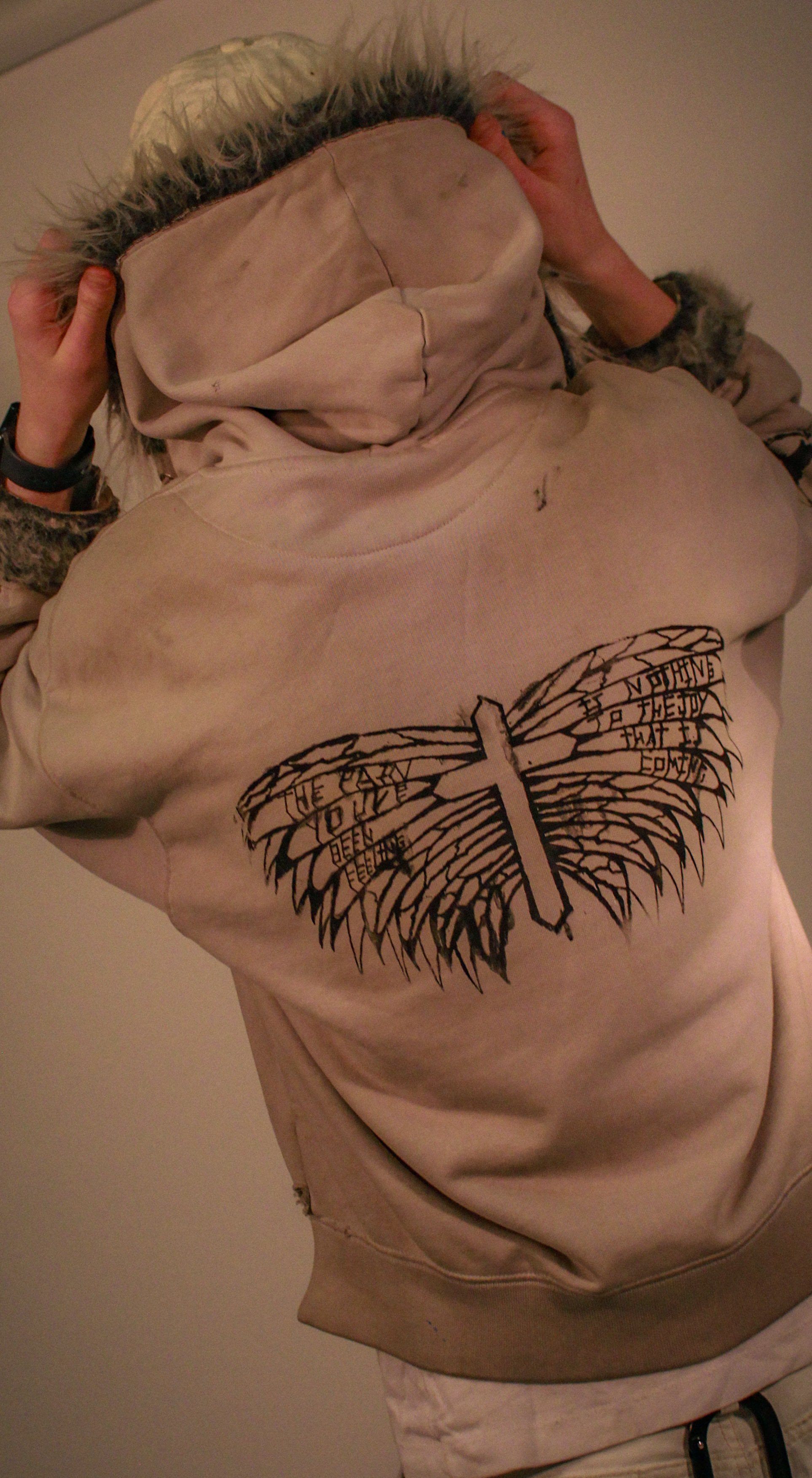 Person holding a hooded sweatshirt with a graphic of a winged cross and text on the back.