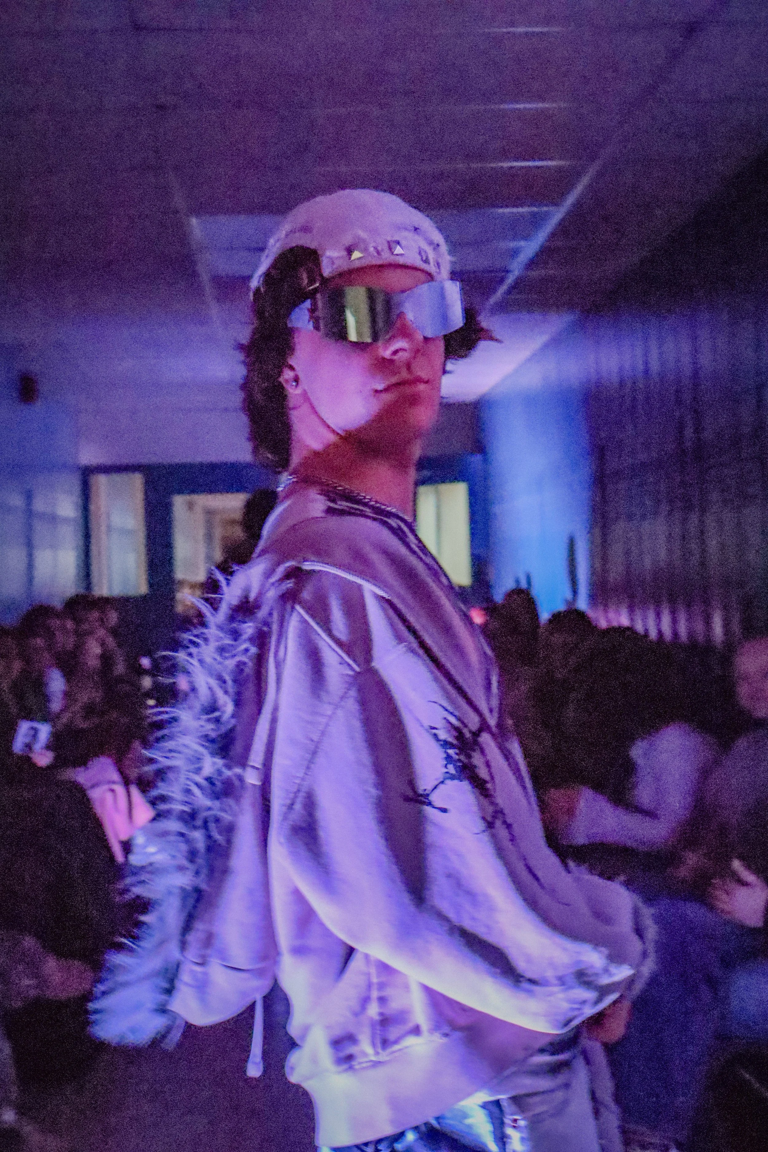 A young person with dark hair wearing a white cap, reflective goggles, and a satin bomber jacket with feather detailing on the sleeves, standing in a dimly lit room with purple and pink lighting.