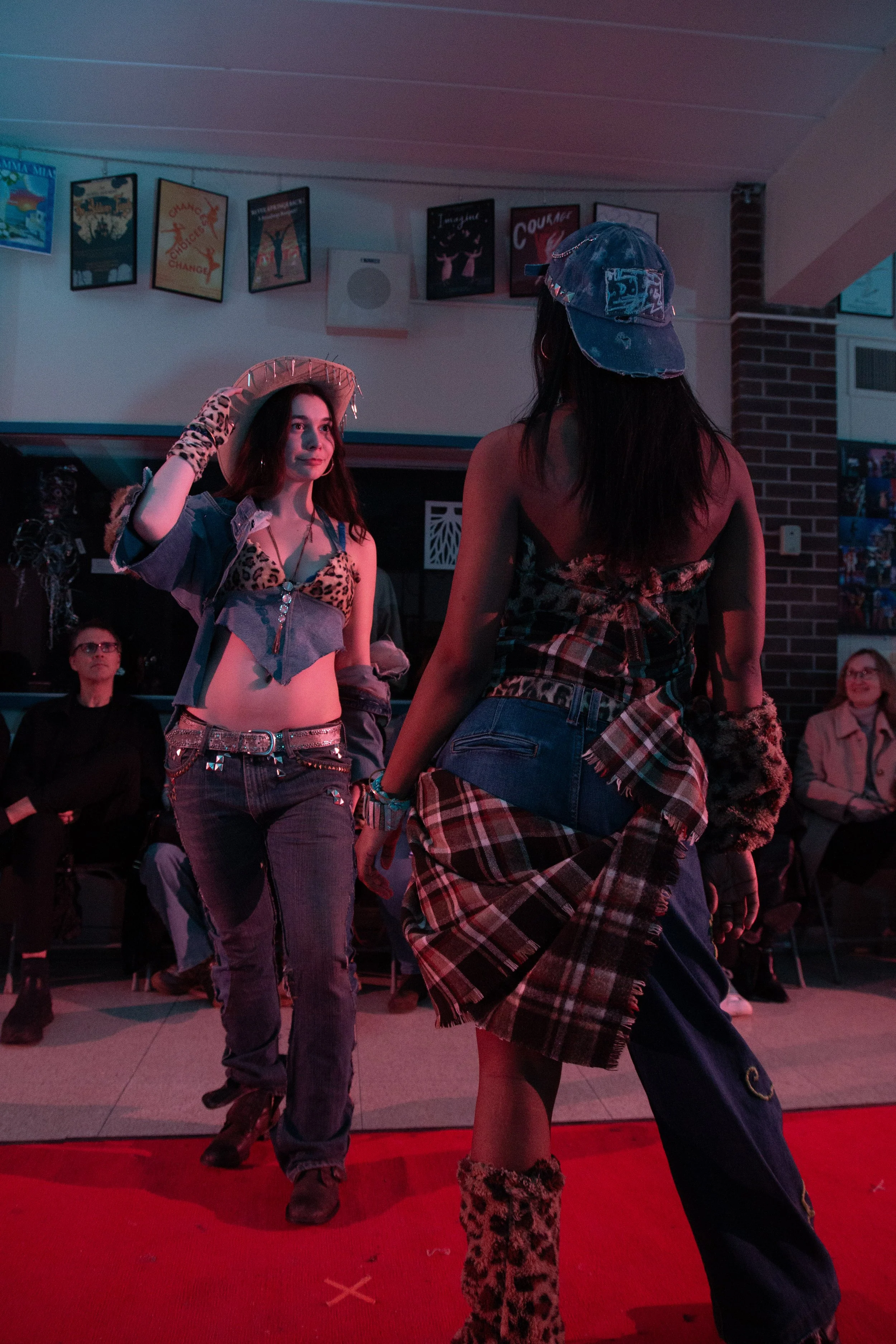 Two women posing on a runway with posters on the wall, one wearing a leopard print top, jeans, gloves, and a hat, the other in a denim hat, plaid skirt, and leopard print boots. Audience seated in the background.