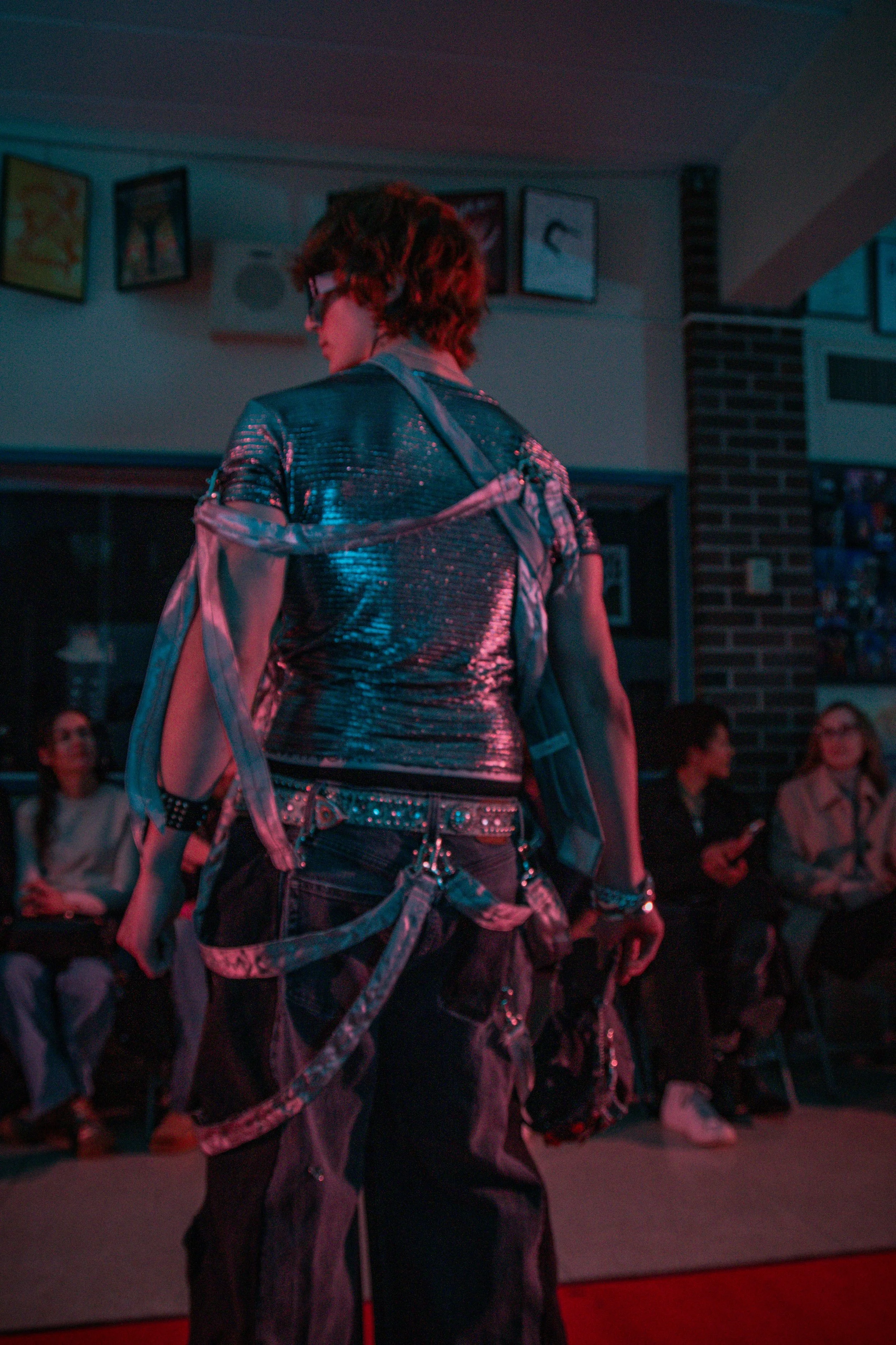 A person with short, curly hair wearing glasses and a shiny, metallic top, with chains and straps hanging from their clothing, is standing in front of an audience in a dimly lit room with framed pictures on the walls.