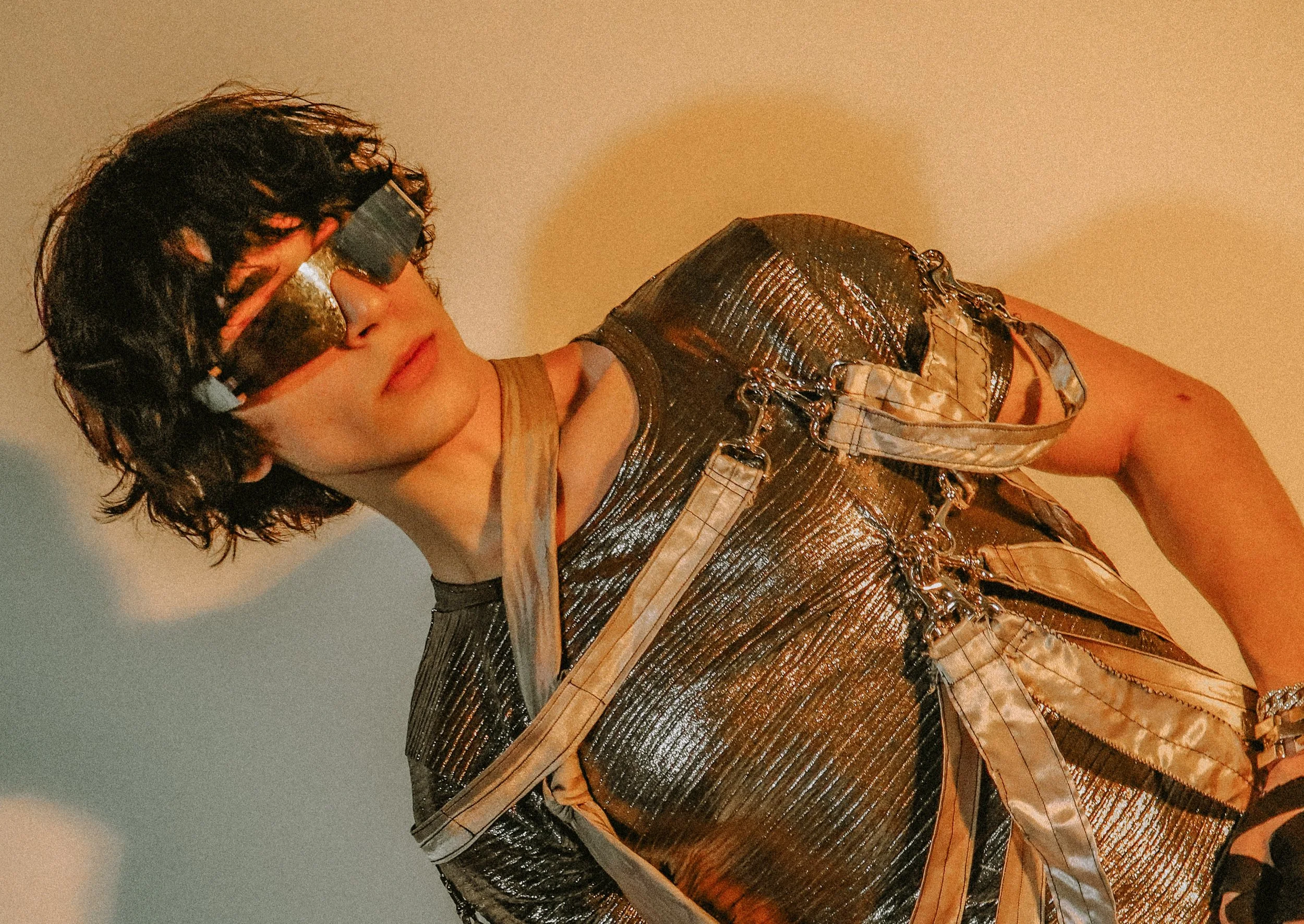 A person with curly dark hair wearing large futuristic silver reflective sunglasses, a metallic silver textured shirt, and a layered outfit with straps, chains, and accessories, standing against a plain background with yellow and blue lighting.