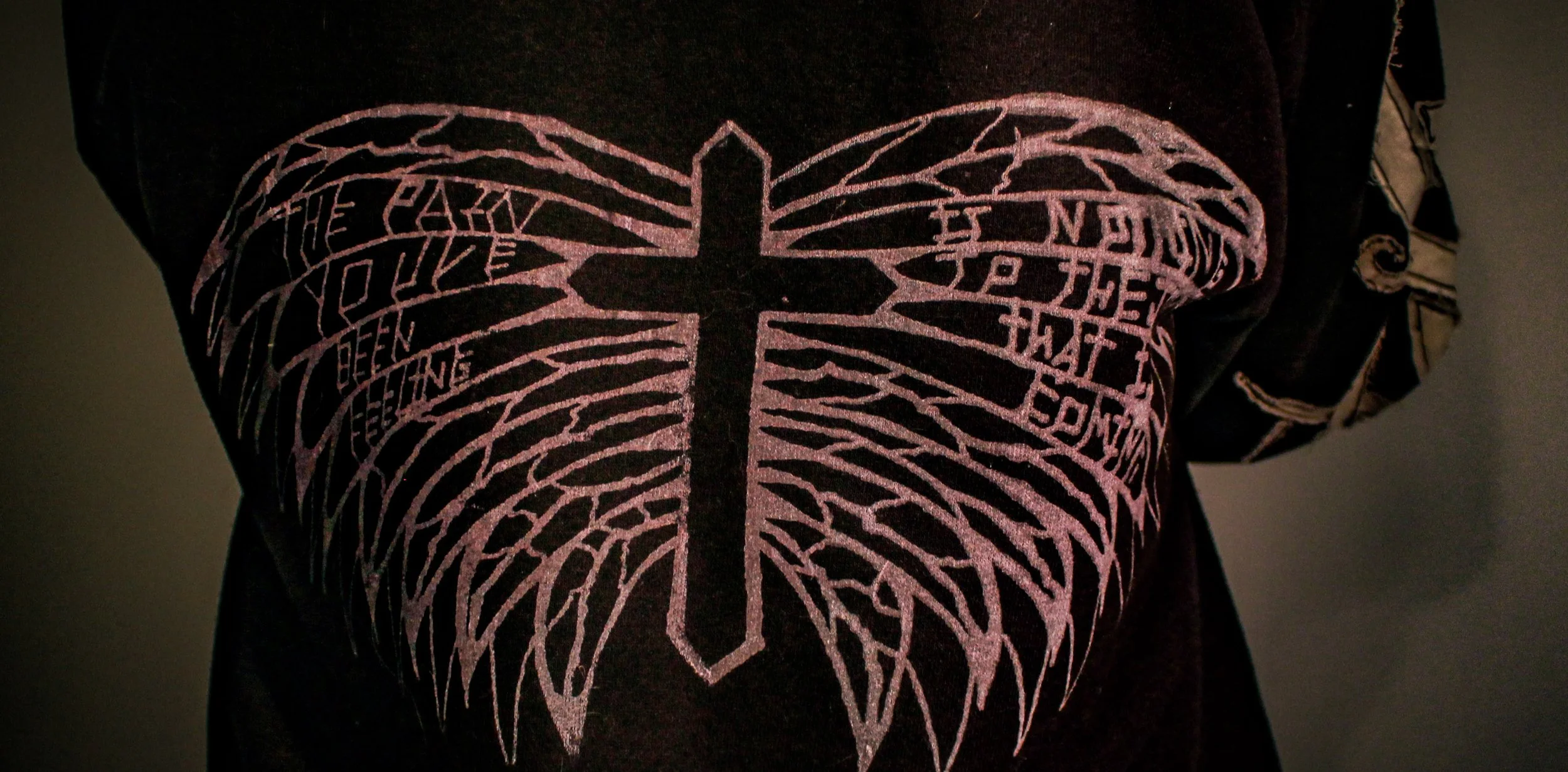 Close-up of a person wearing a black shirt with a white graphic of a cross with wings that have words inside of them.