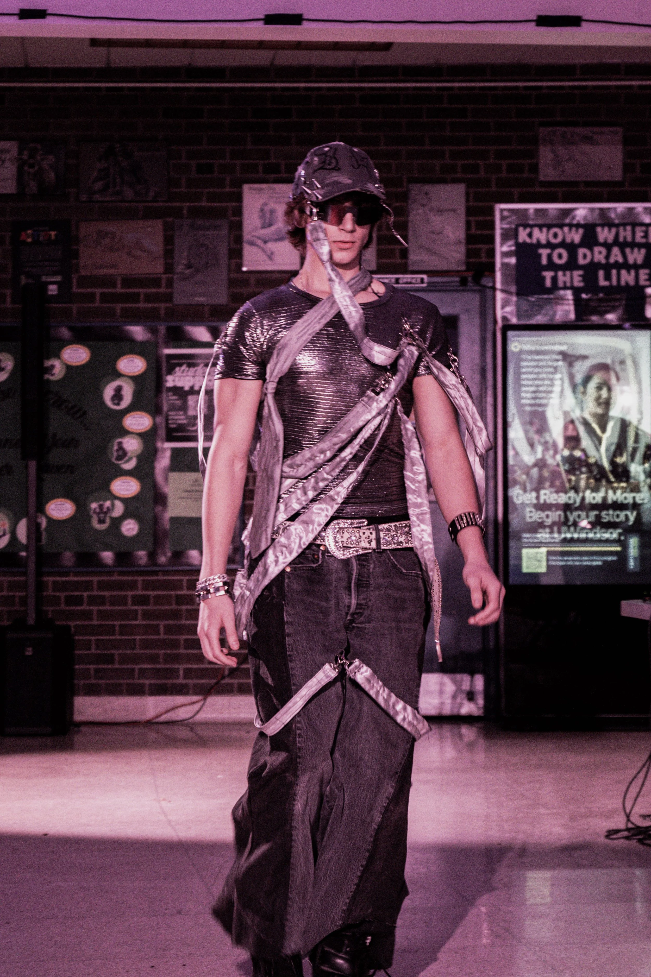 A person dressed in a shiny, metallic silver outfit with a matching hat, glasses, and gloves, walks on a runway in front of an audience, with purple lighting. Behind them are posters and a brick wall.