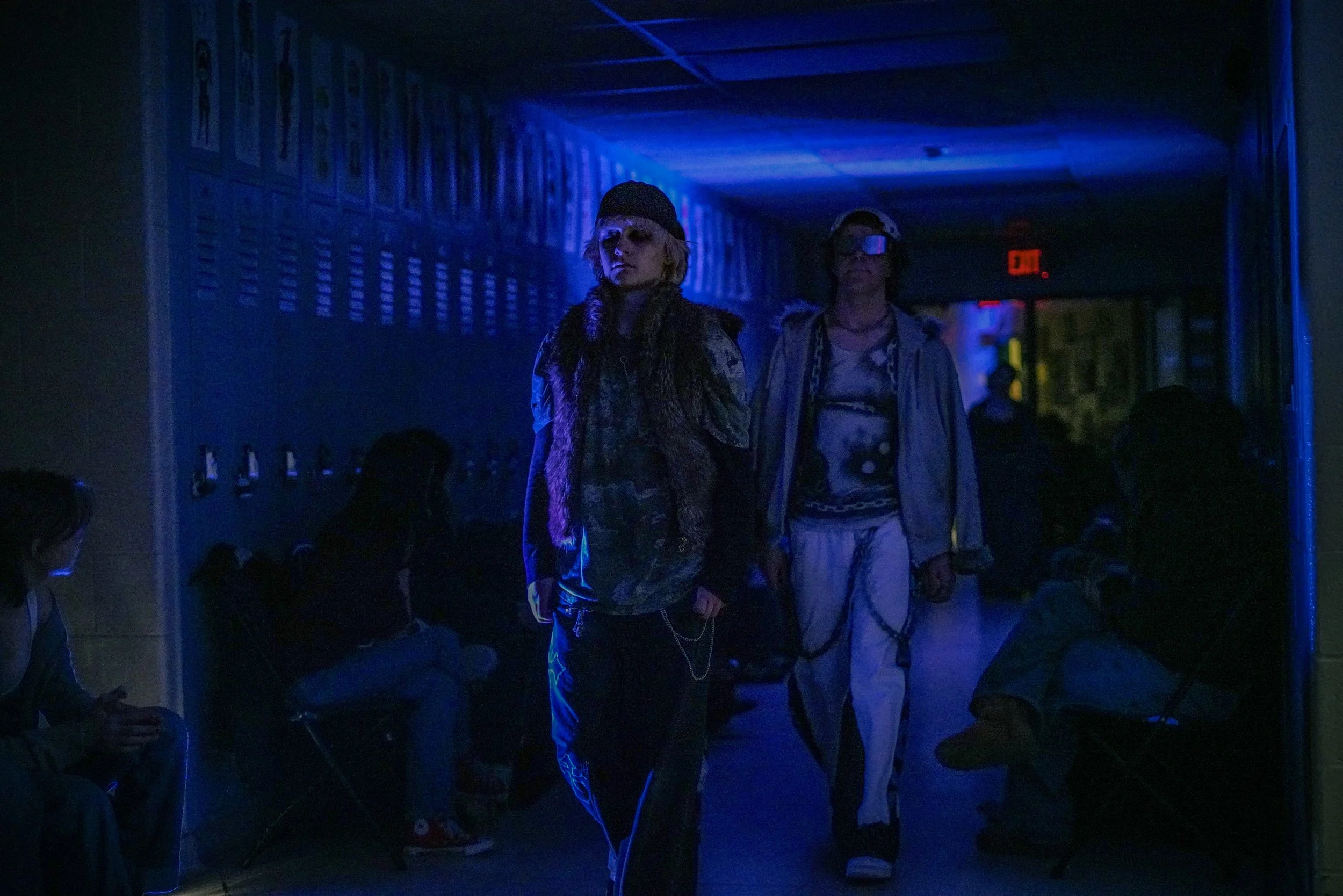 A person dressed in streetwear with chains, glasses, and a cap, walking in a dimly lit indoor space with blue lighting and other people in the background.