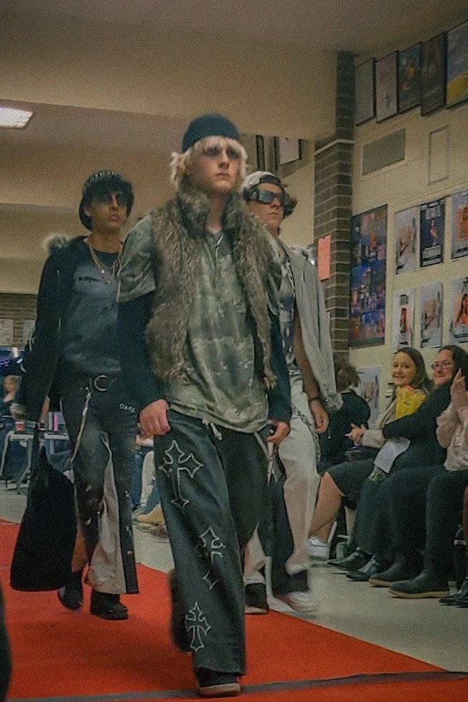 Three young men walking on a red carpet, dressed in fashionable streetwear, at a fashion event, with seated audience members watching and smiling in the background.