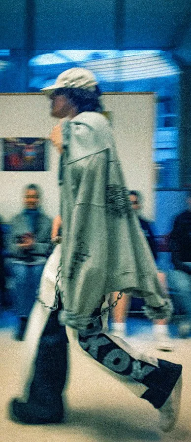 A person walking through an airport terminal wearing a large gray hoodie, black pants with white lettering, and a beige baseball cap, with other travelers seated in the background.