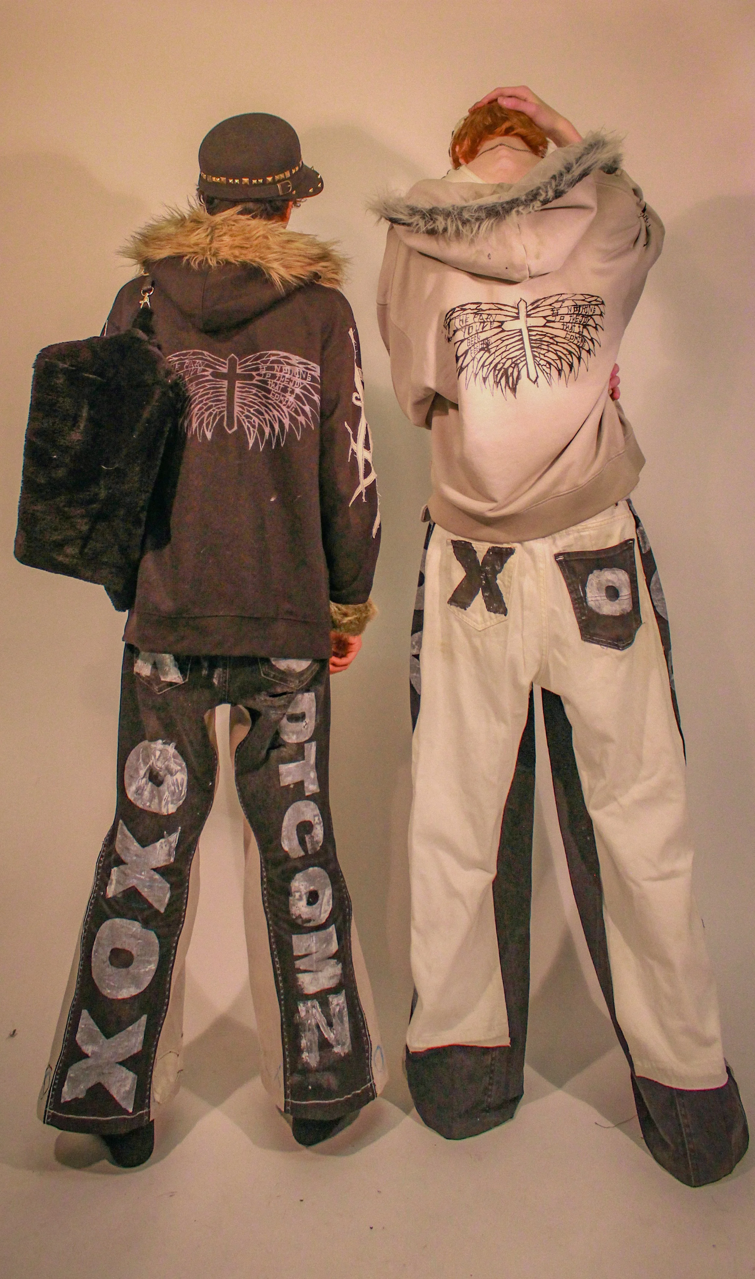 Two people wearing oversized clothing with cross and wing designs on the back, standing against a plain wall.