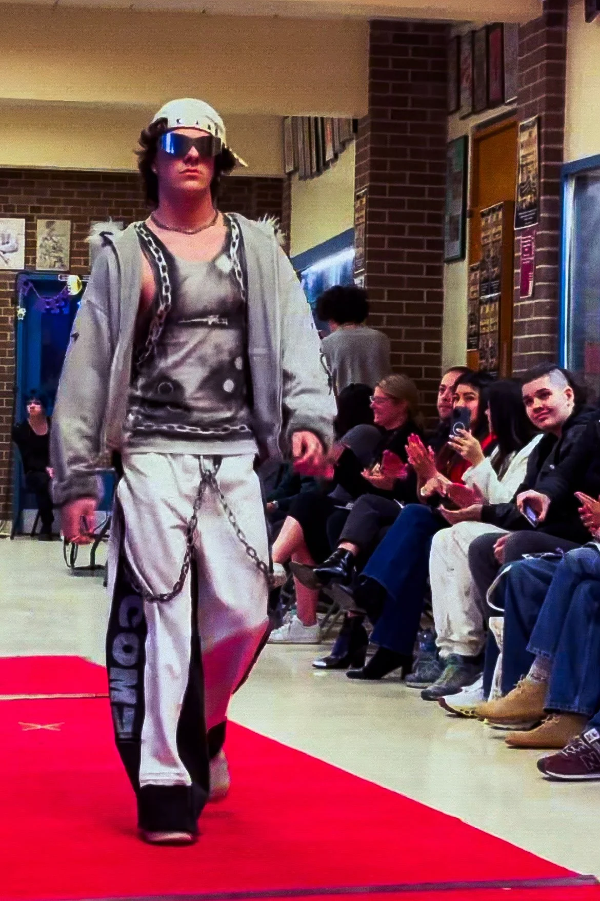 A young male model walking on a red carpet runway at a fashion show. He is wearing sunglasses, a backward cap, a stylish tank top, and baggy pants with chains. Spectators are seated on the side clapping and watching.