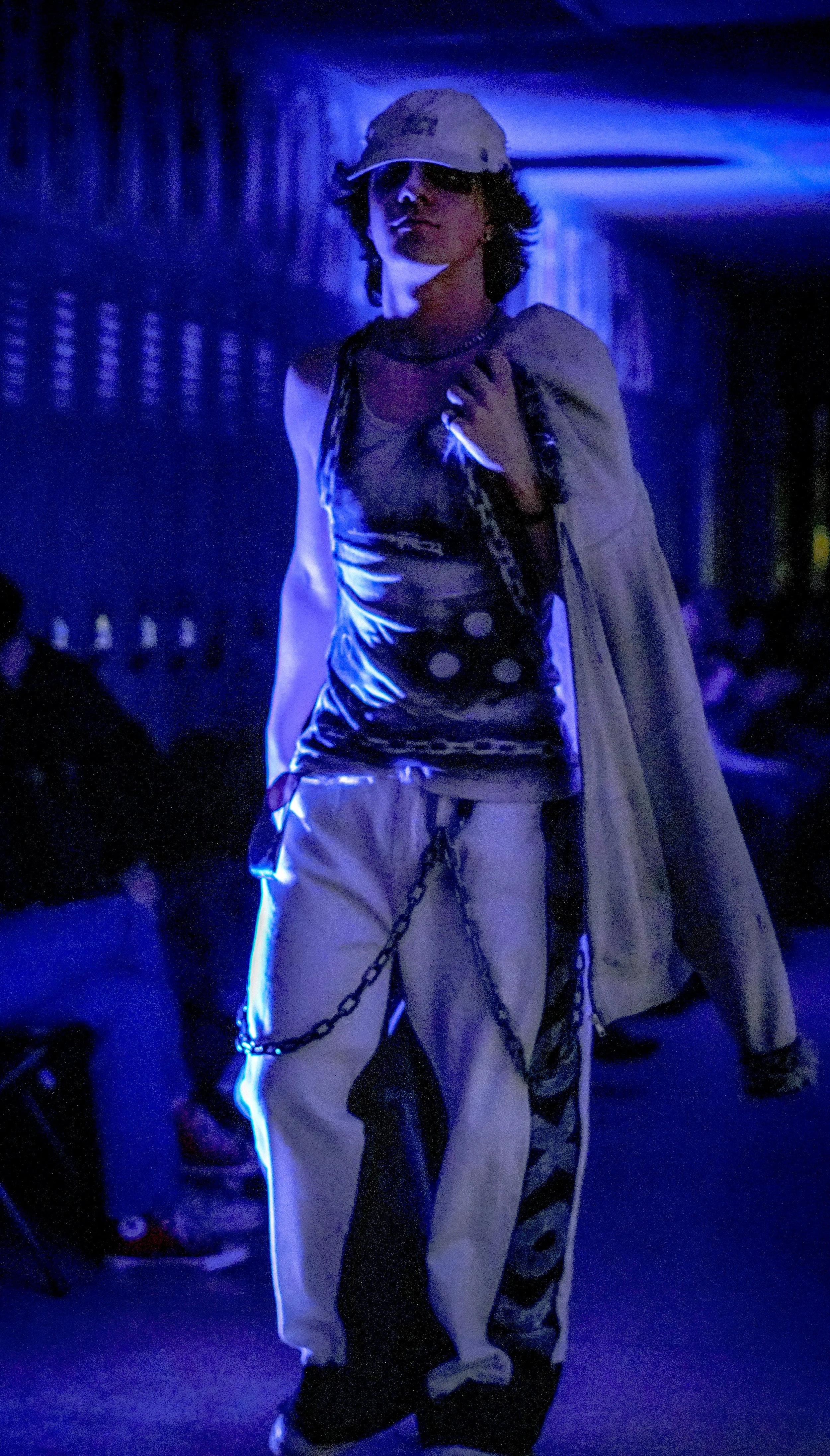 A person dressed in streetwear with chains, glasses, and a cap, walking in a dimly lit indoor space with blue lighting and other people in the background.