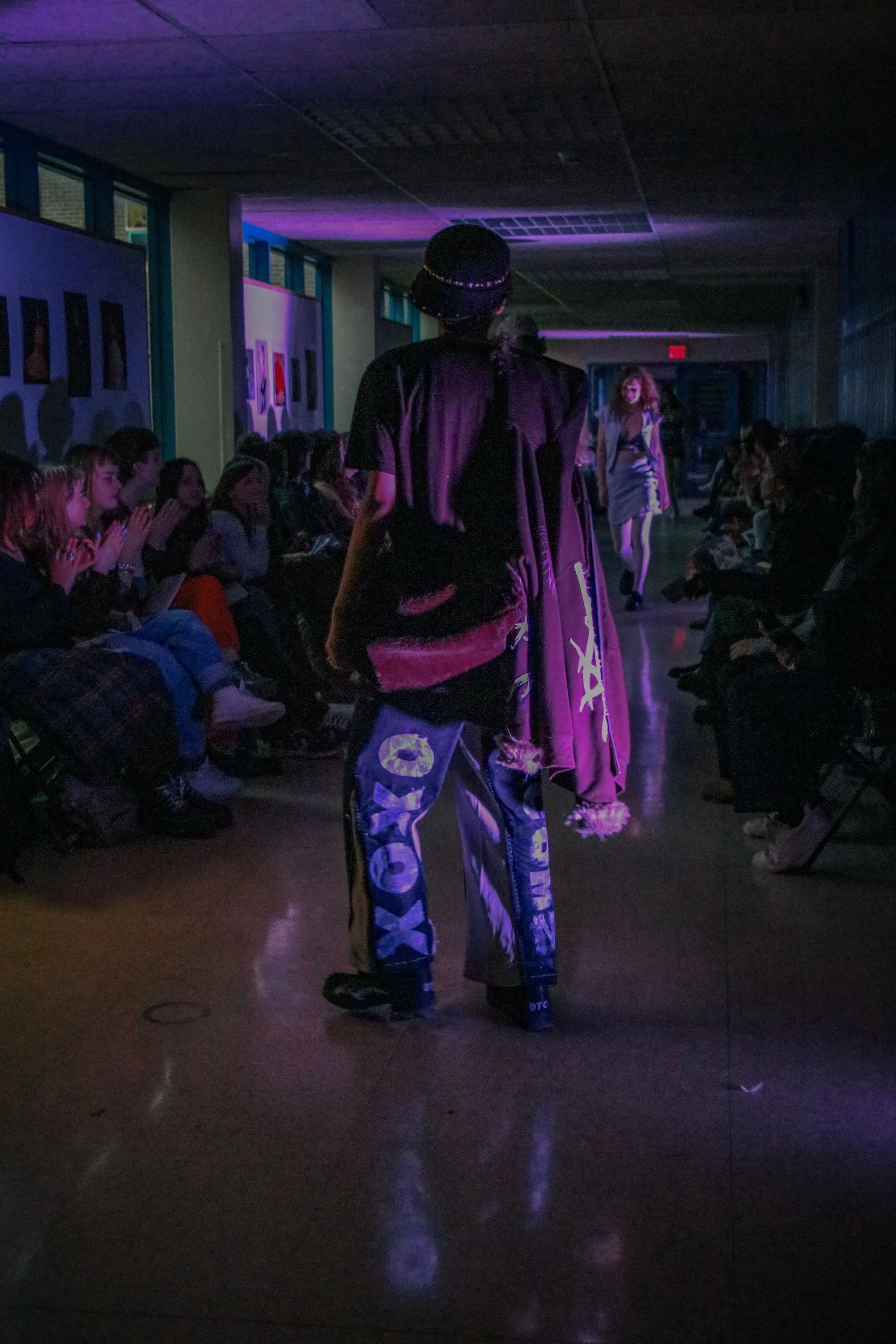 Person walking down a dark runway with purple lighting, audience seated along the sides, and artwork displayed on the walls.