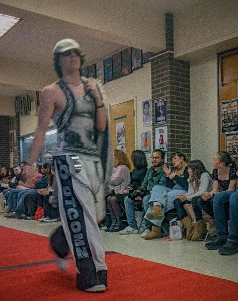 A woman walking on a runway during a fashion show, with an audience seated along the sides of the runway watching.