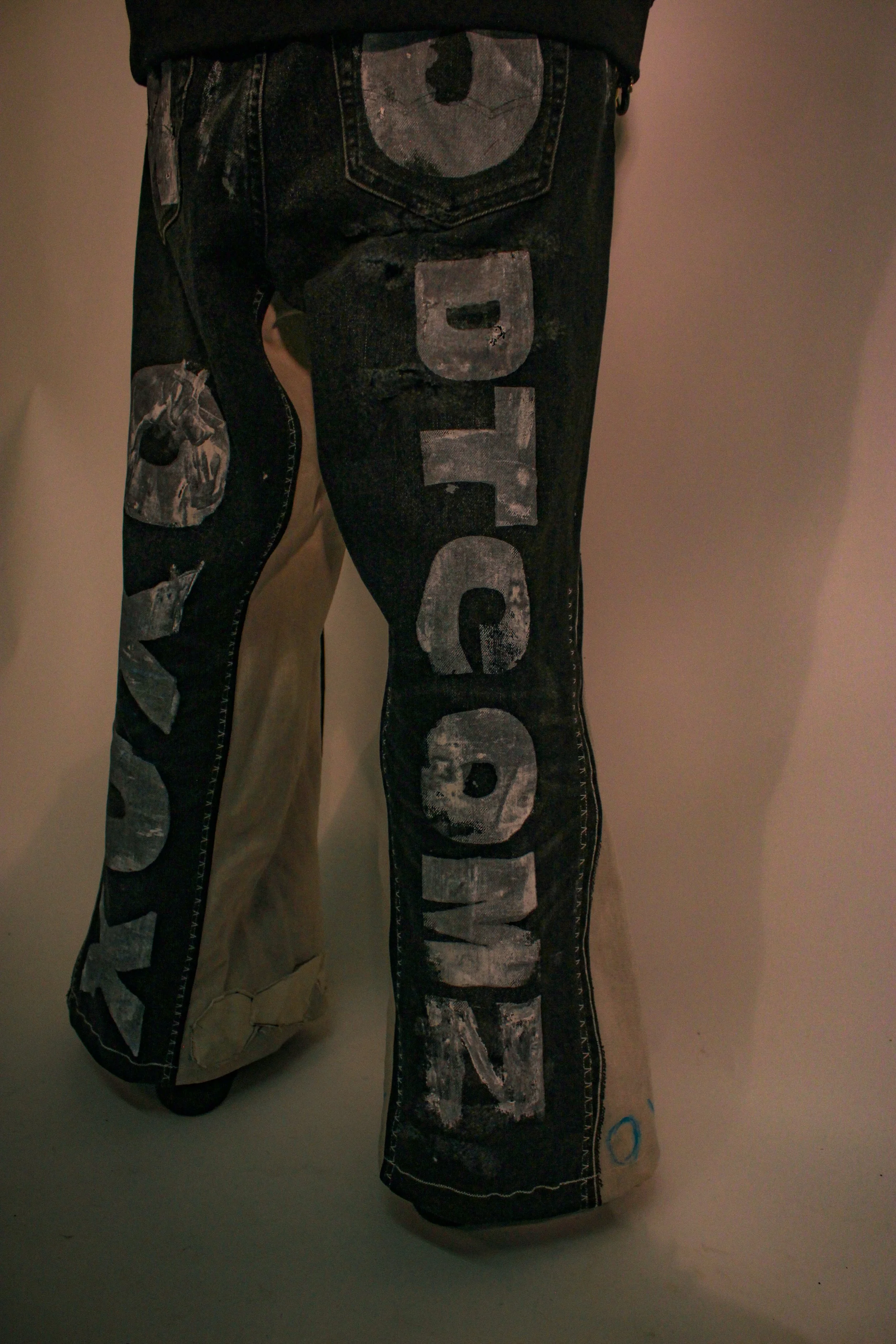 Close-up of a person wearing black jeans with large, faded gray letters spelling 'Dior' down the right leg, paired with beige pants.