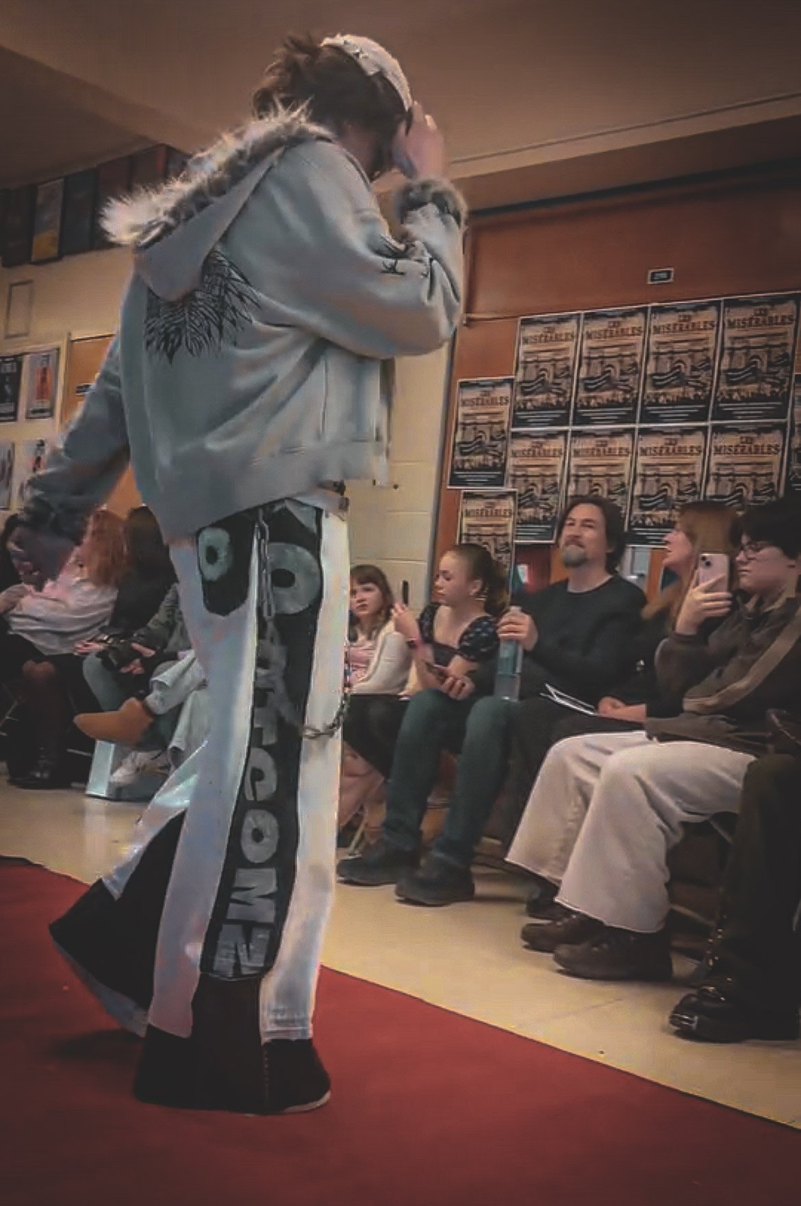 A young male model walking on a red carpet runway at a fashion show. He is wearing sunglasses, a backward cap, a stylish tank top, and baggy pants with chains. Spectators are seated on the side clapping and watching.