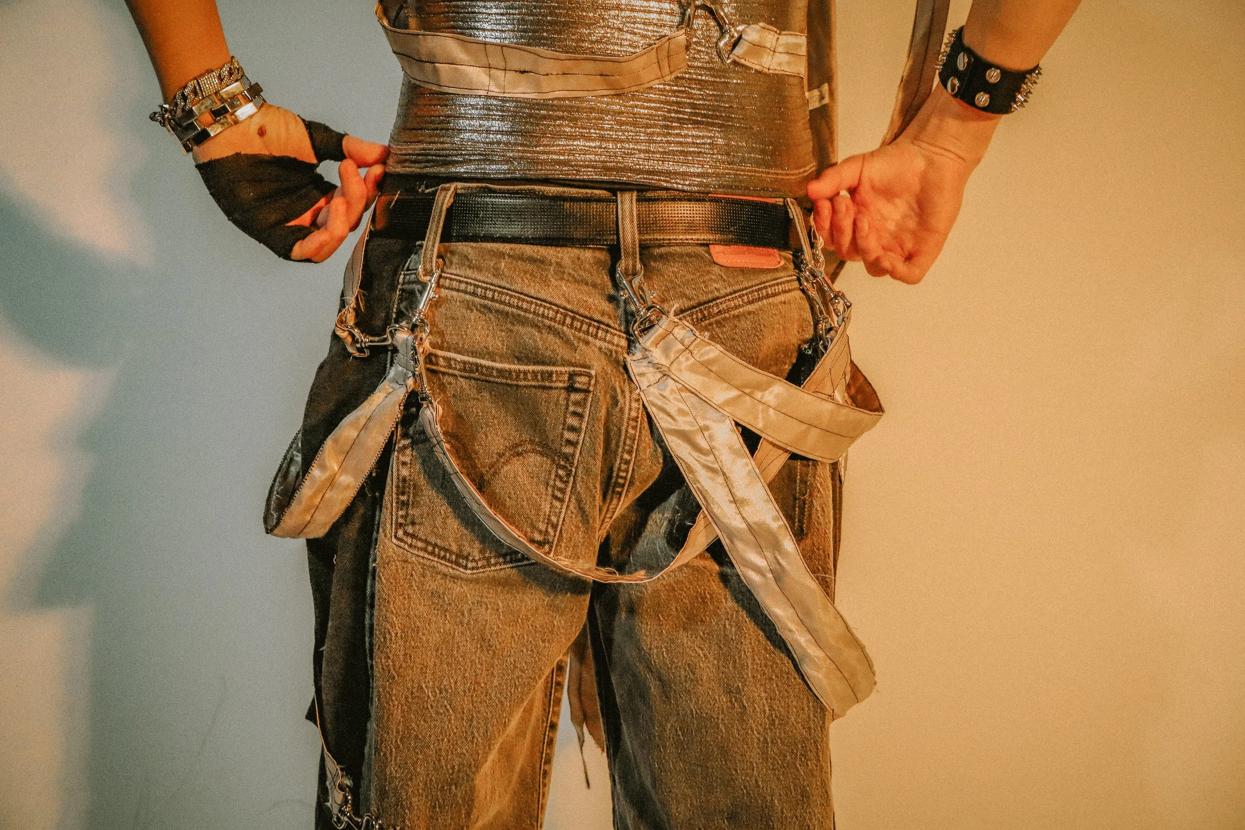 Close-up of a person's midsection wearing distressed jeans with a black belt, a metallic silver oversized top, and accessories including straps, a black glove, and a studded wristband.