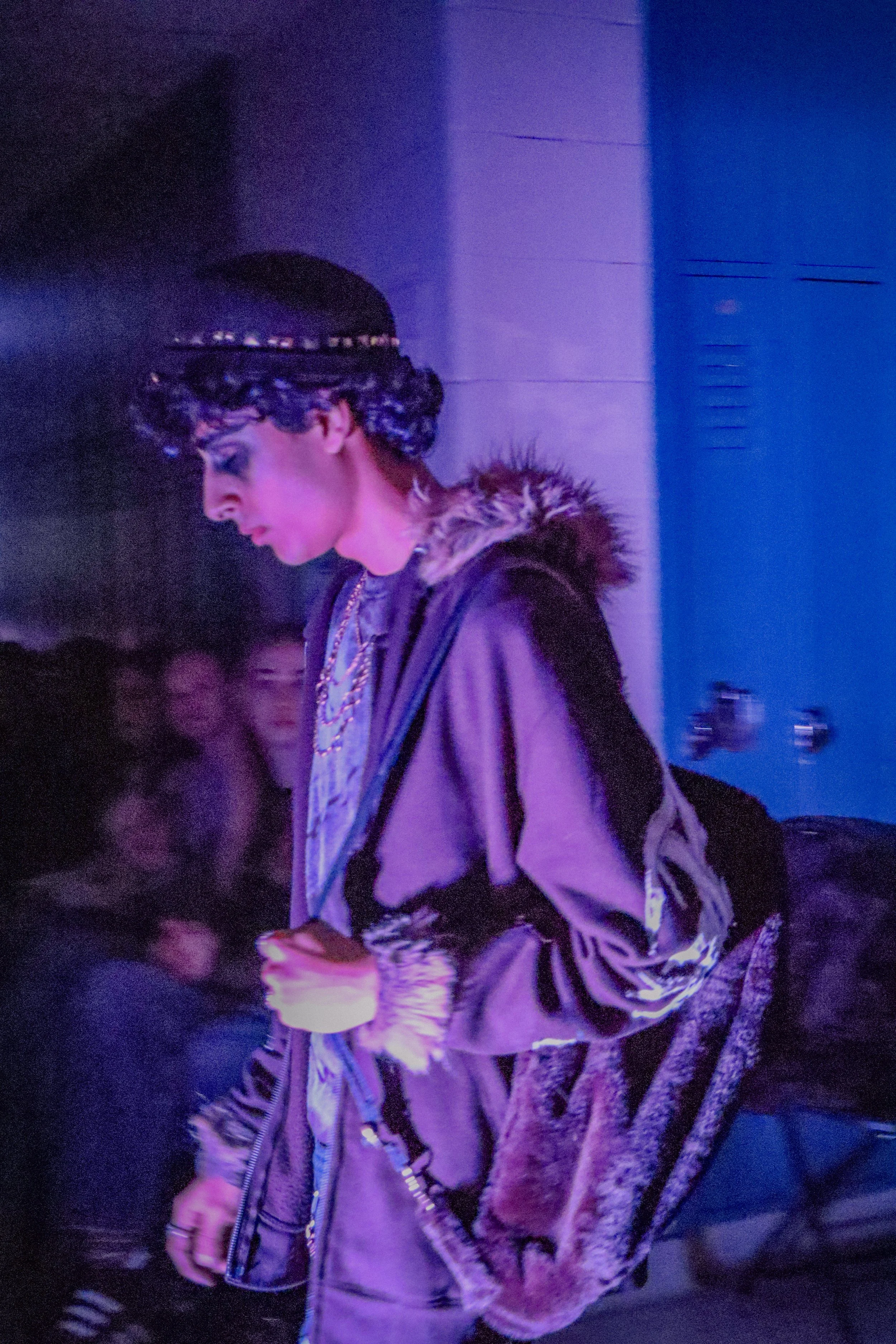 A person with curly hair wearing a black beanie with embellishments, a fur-lined jacket, and carrying a patterned backpack, walking in a dimly lit environment with purple lighting and blurred background.