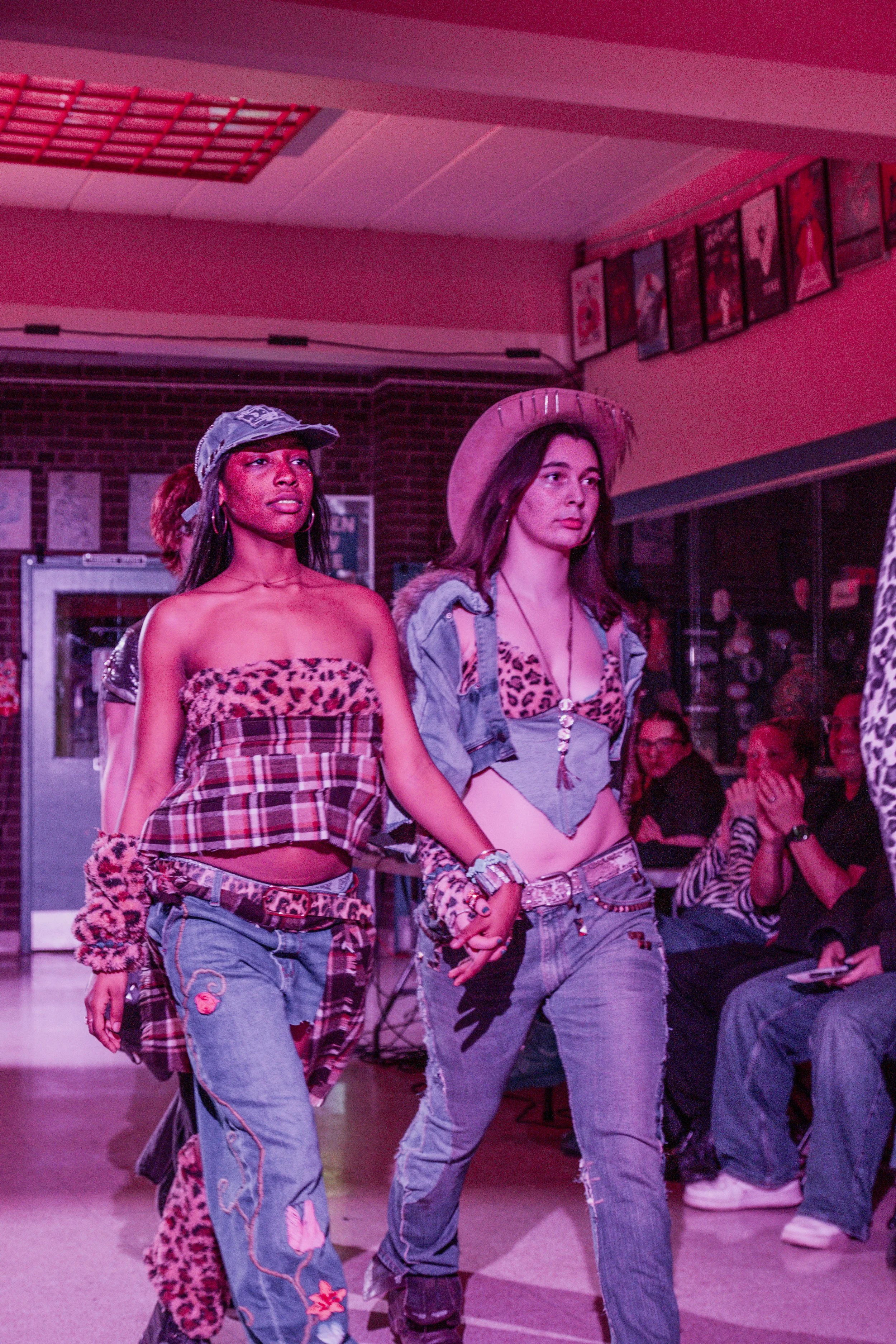 Two women walking in a fashion show, wearing punk-inspired outfits with leopard print tops, denim jeans with patches, and accessories, in front of an audience.