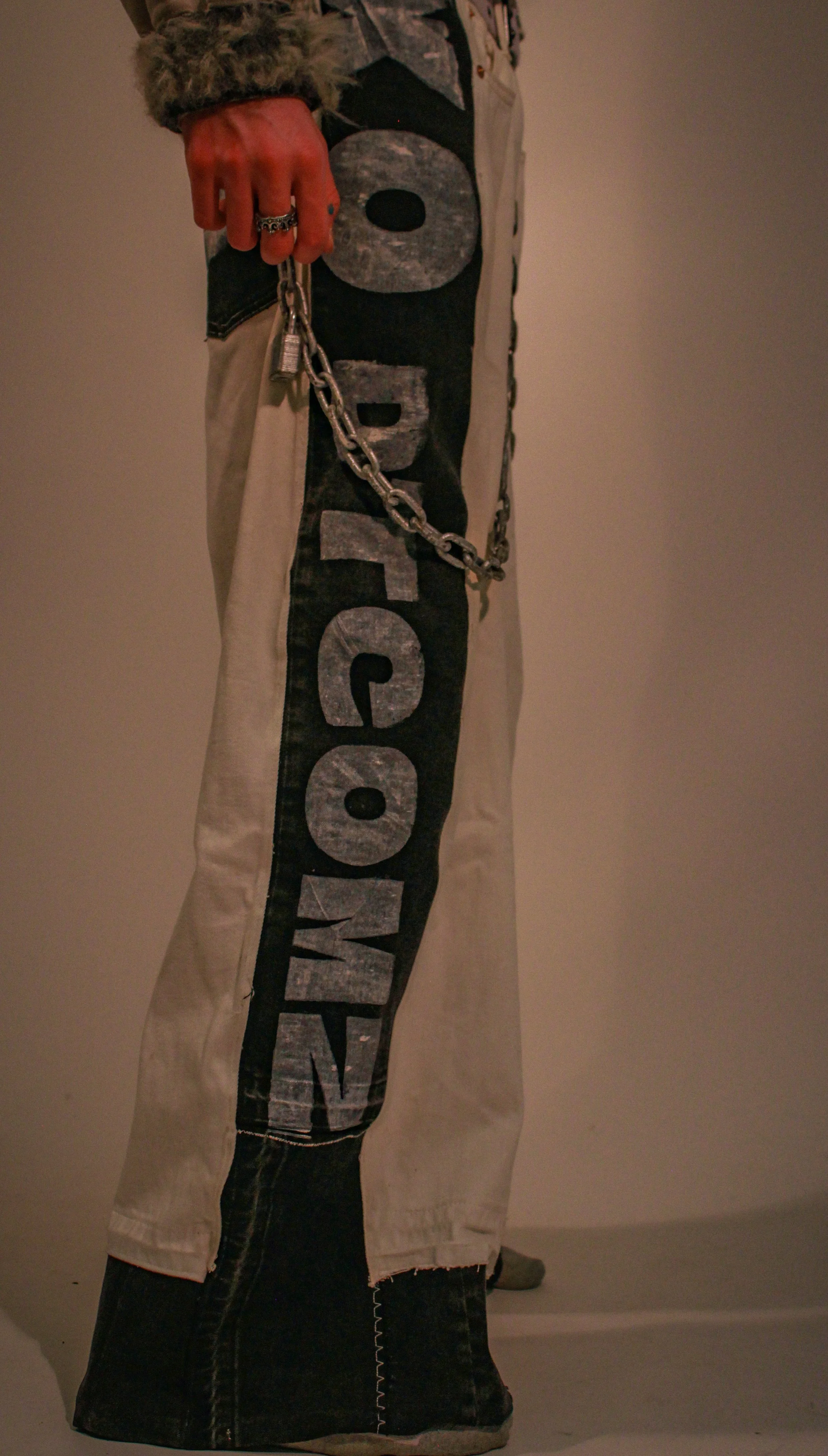 Close-up of a person wearing beige pants with large black lettering reading 'DTCOMZ' on the side, with a chain attached to their belt loops. The chain has a padlock on it.
