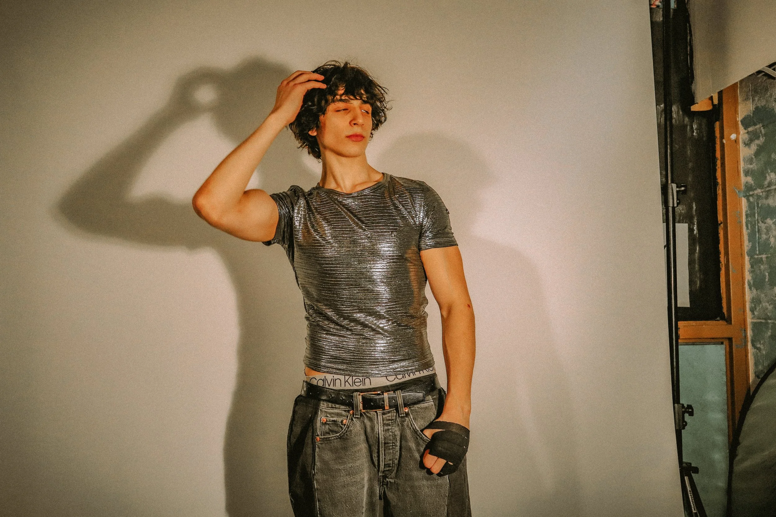 A young person with curly dark hair posing in a photoshoot studio, wearing a metallic-patterned t-shirt, black jeans with a Calvin Klein waistband, and a black fingerless glove, adjusting their hair.