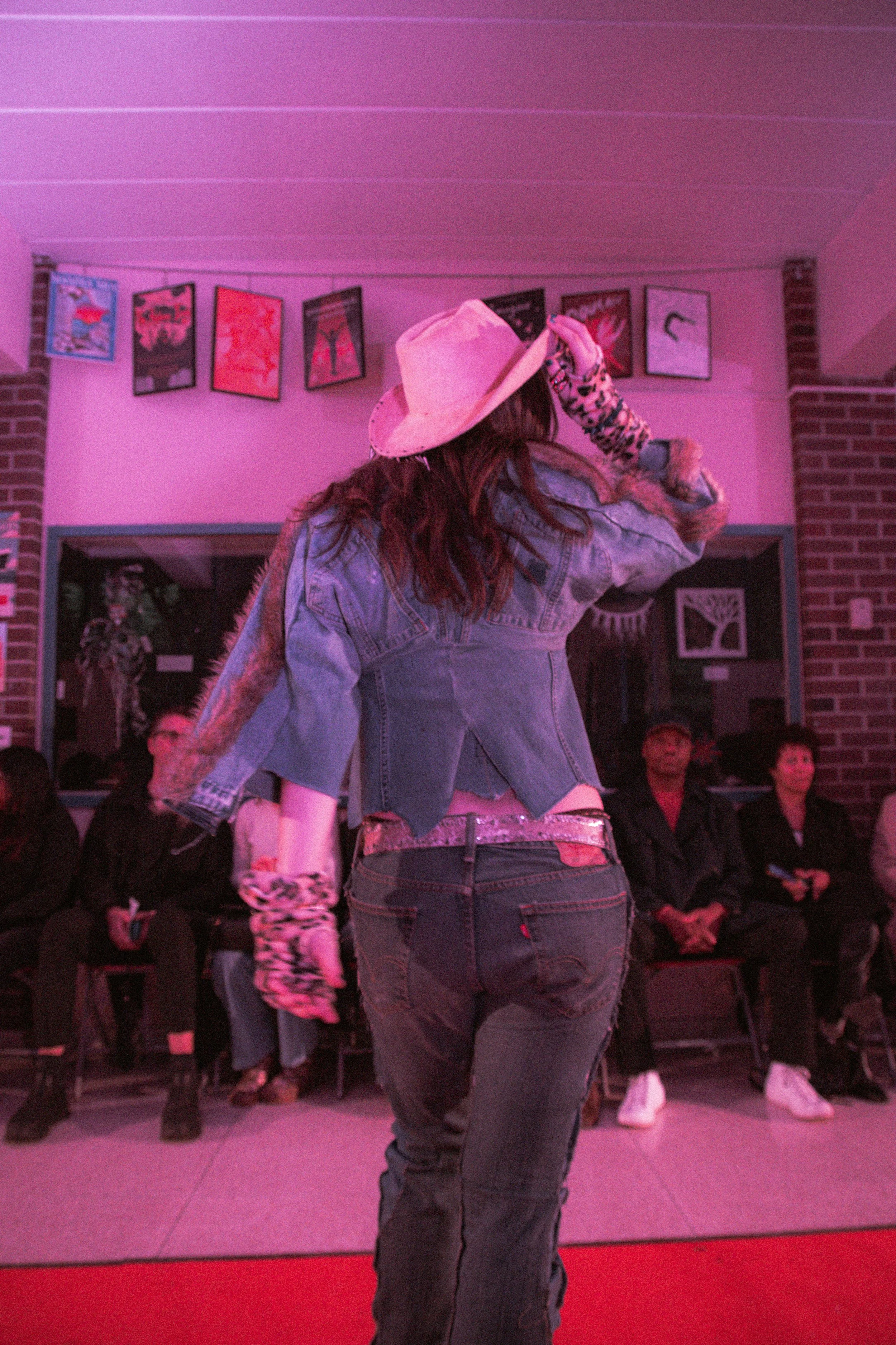 A person wearing a denim jacket and jeans, with a pink hat and patterned gloves, dancing on a red carpet in front of an audience, with framed wall art in the background.