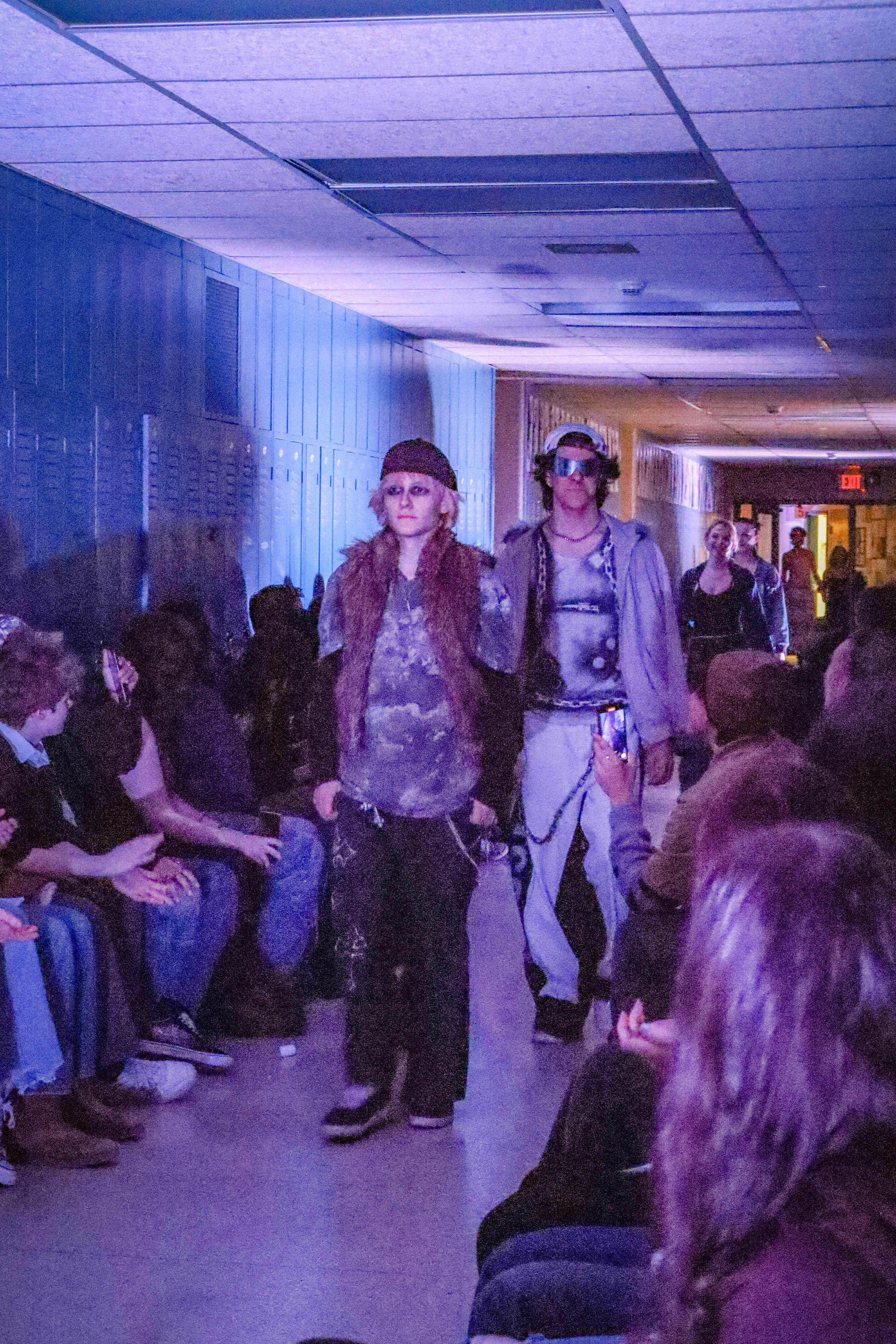 People walking through a dimly lit corridor with purple lighting, some seated along the sides, and others standing or walking. The ceiling is covered with panels, some of which are missing or discolored. The scene appears to be at a public event or g