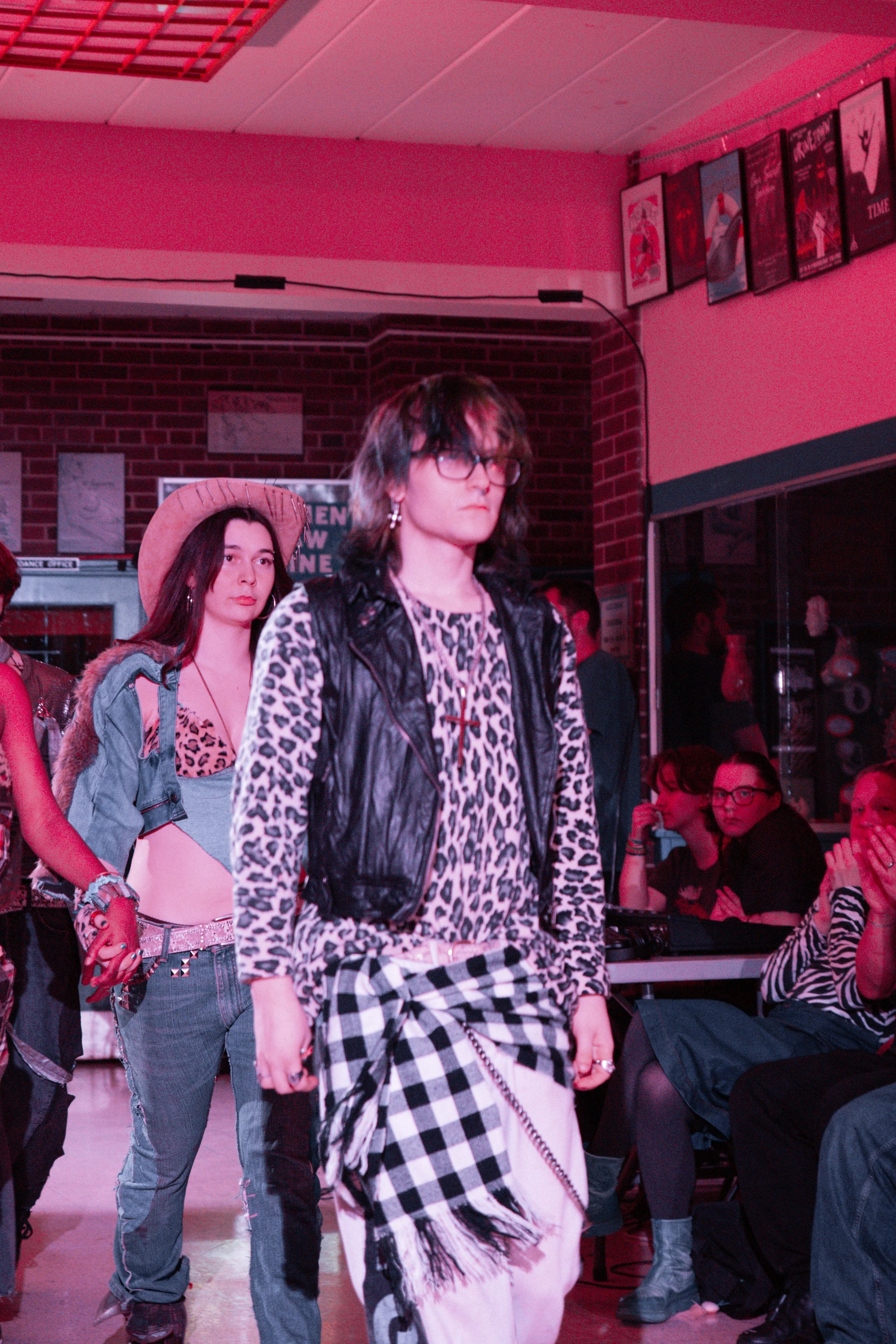 People walking on a runway with pink lighting, including two women in the foreground with edgy fashion, one with leopard print clothing and glasses, the other in a styled hat and denim. Others are sitting and standing in the background.