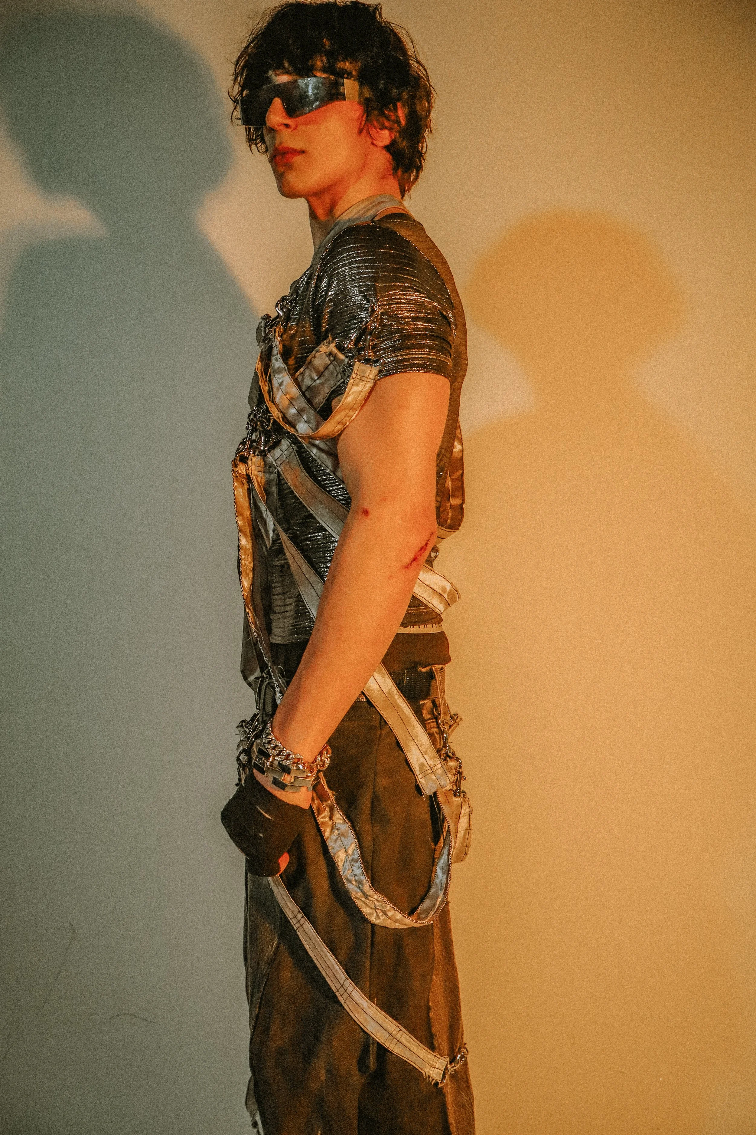 A person with curly dark hair wearing large futuristic silver reflective sunglasses, a metallic silver textured shirt, and a layered outfit with straps, chains, and accessories, standing against a plain background with yellow and blue lighting.