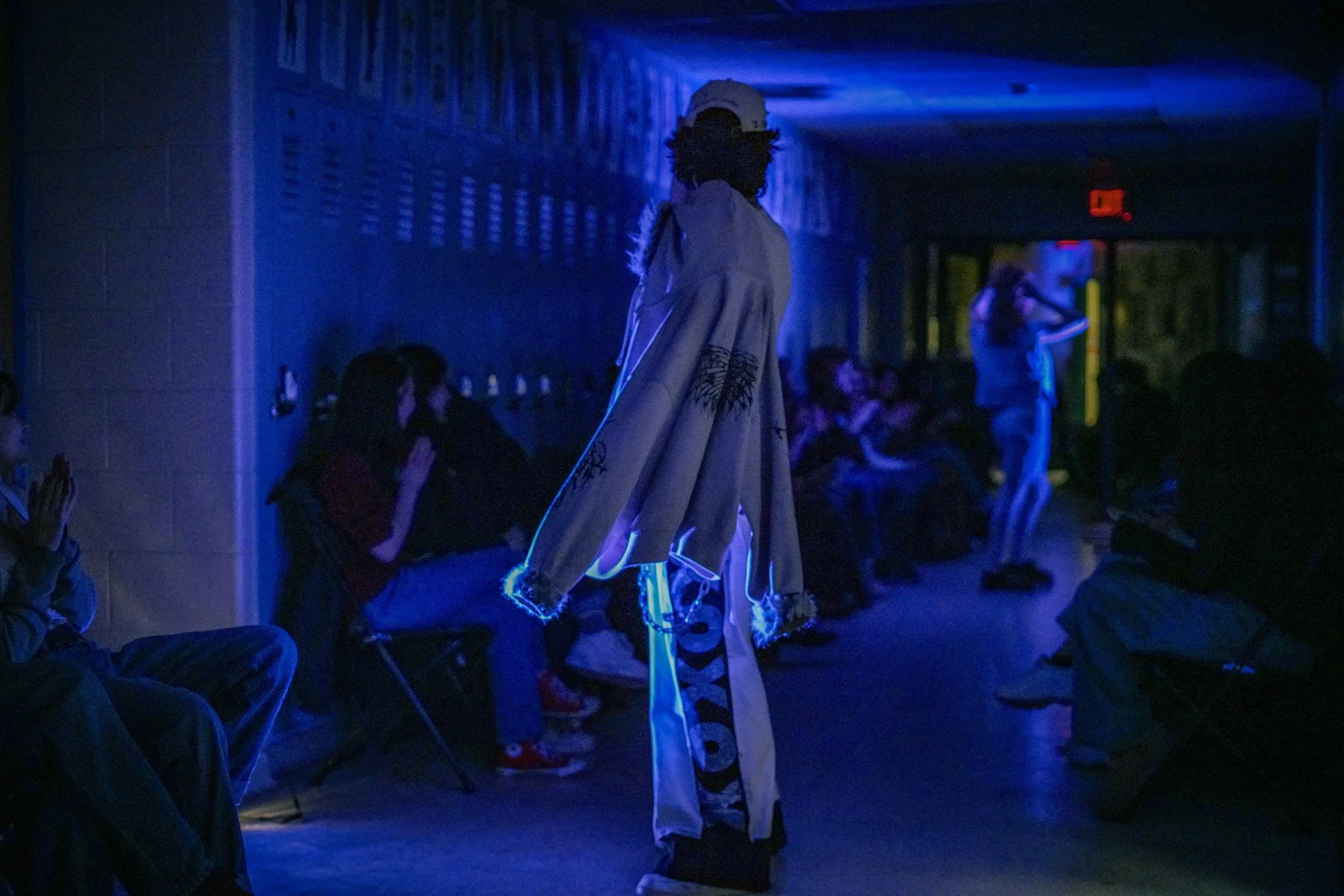 A person dressed in streetwear with chains, glasses, and a cap, walking in a dimly lit indoor space with blue lighting and other people in the background.