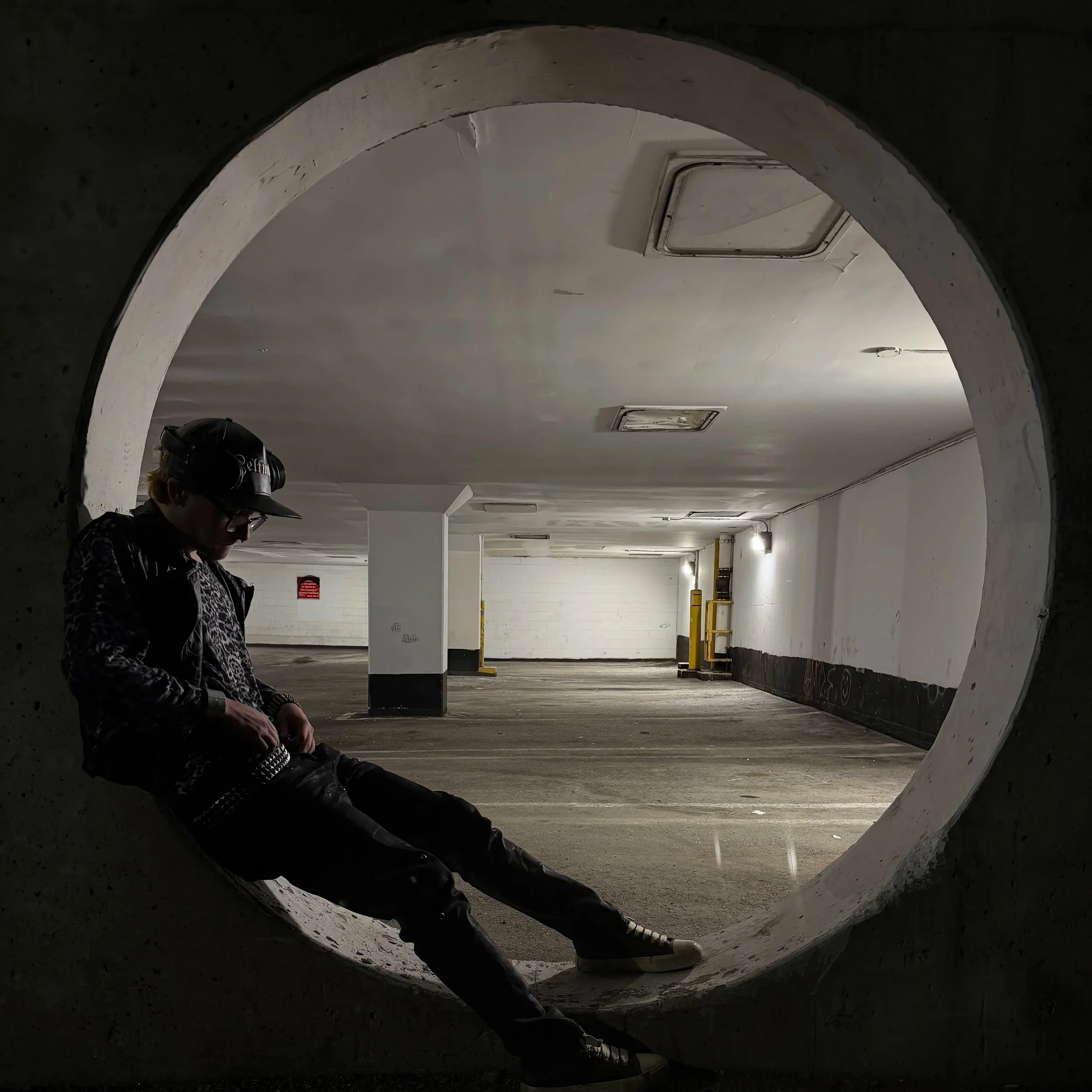 Person wearing a hat  and glasses sitting on the ground in a parking garage, seen through a circular opening in a concrete wall, with ceiling vents and parking lot elements in the background.