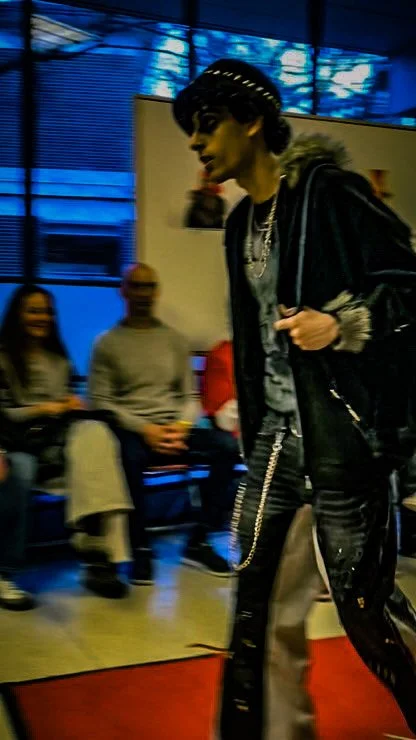 A person with black hair in a short hairstyle, wearing a black leather jacket, gray shirt, and black pants, walking inside a room with a red carpet. In the background, seated people are visible, some smiling, and there are large windows with blue cur