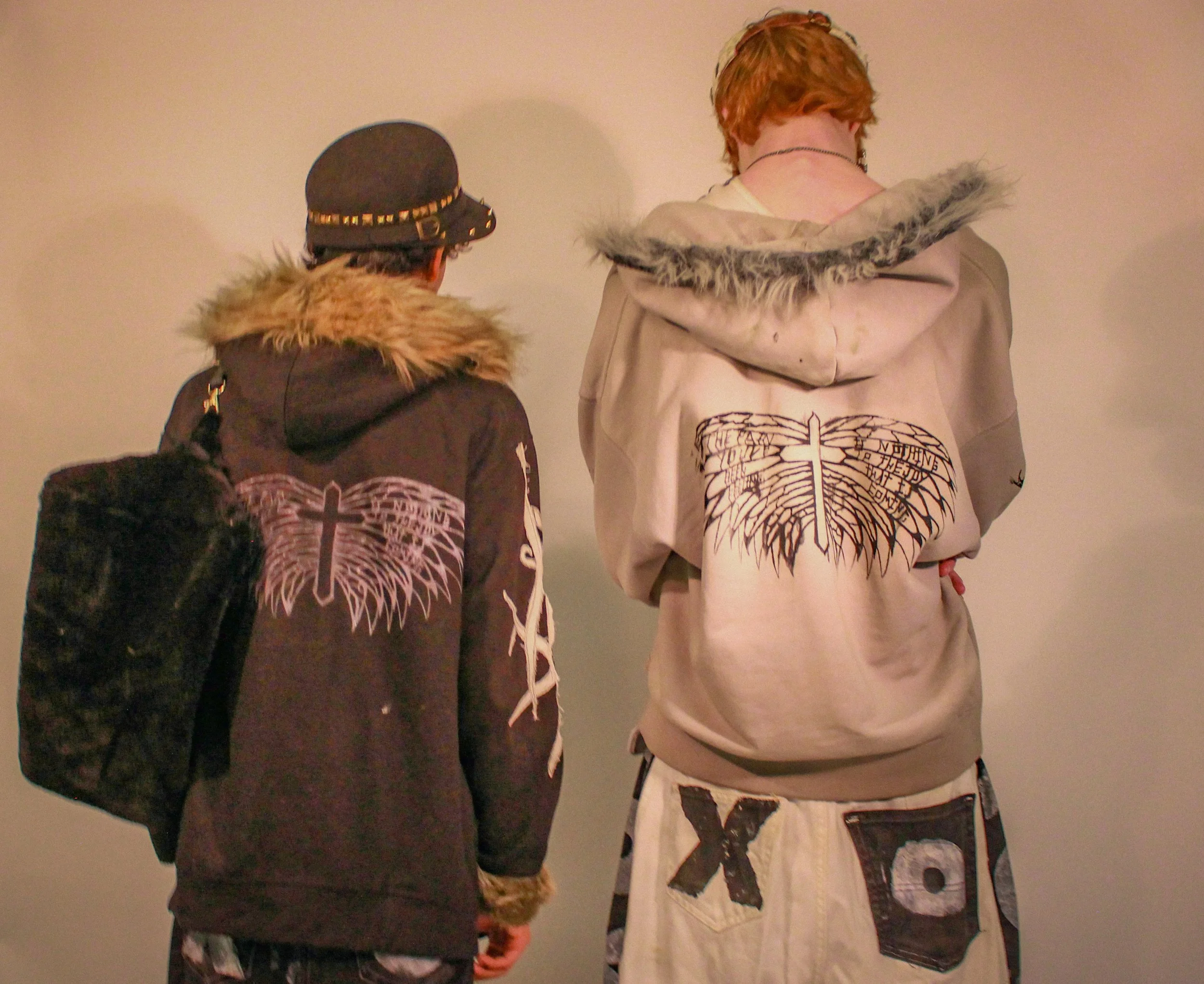 Two individuals standing against a plain wall, viewed from behind. The person on the left wears a black hoodie with a graphic design of a winged cross, a fur-trimmed hood, and a black hat with studs and a small chain. The person on the right has shor