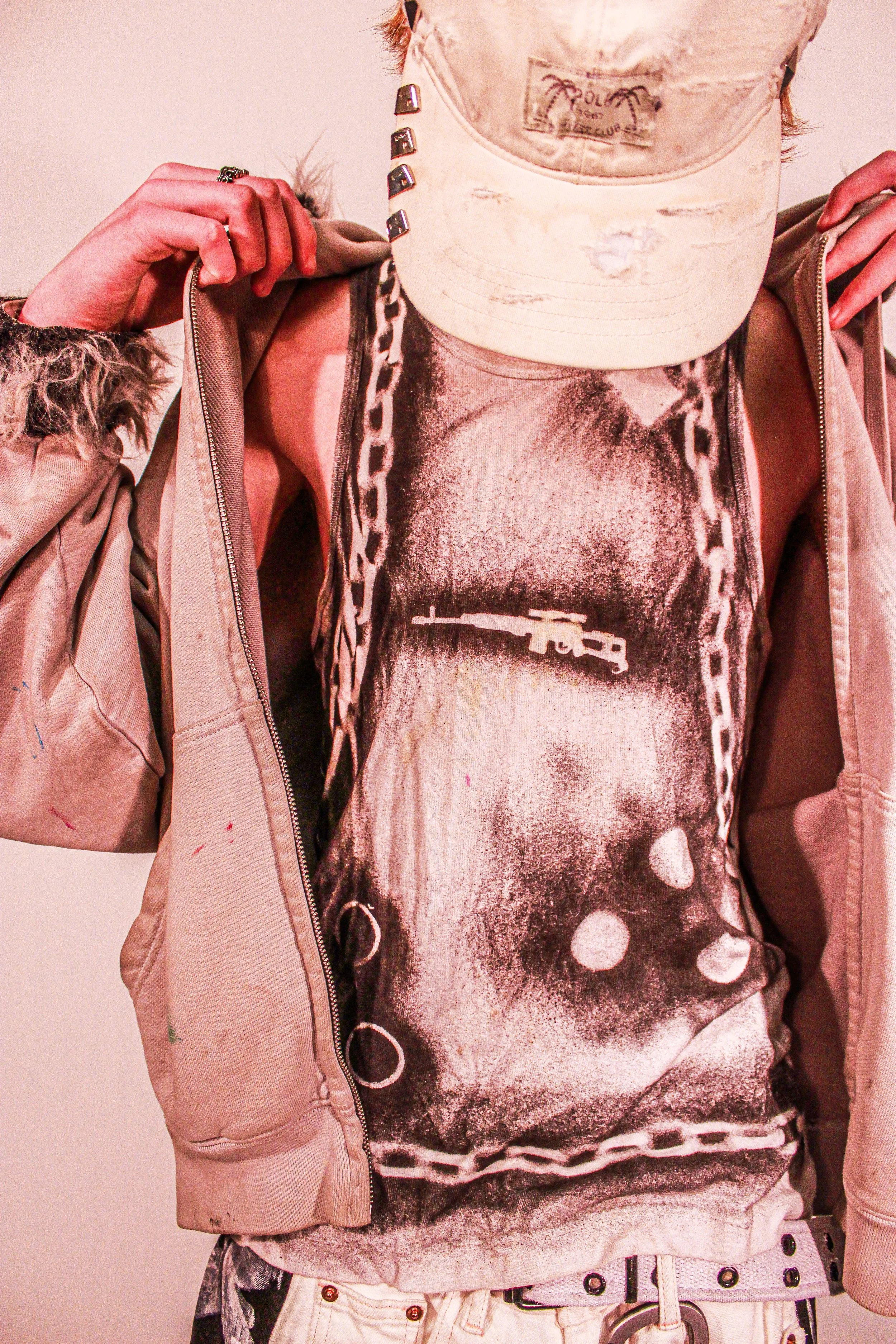 Close-up of a person wearing a white baseball cap, a white graphic tank top, a white sweater with a distressed fur collar, and high-waisted pants with a belt and metal eyelets. The individual is adjusting the collar of the jacket with both hands.