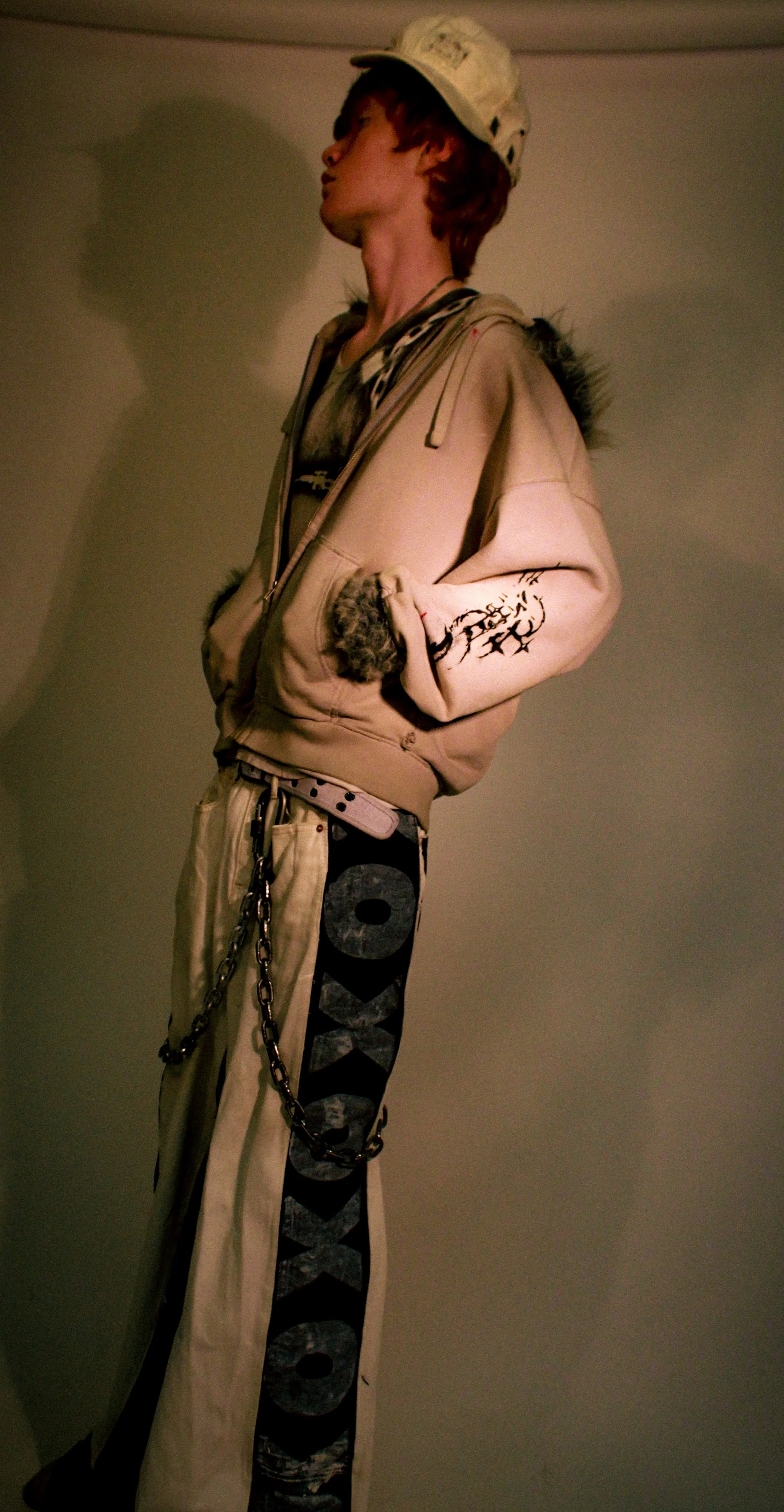 A mannequin wearing a beige hoodie with fur-lined pockets, black pants with large gray lettering, a white belt, and chains hanging from the waist. The mannequin has a white cap and styled brown hair.
