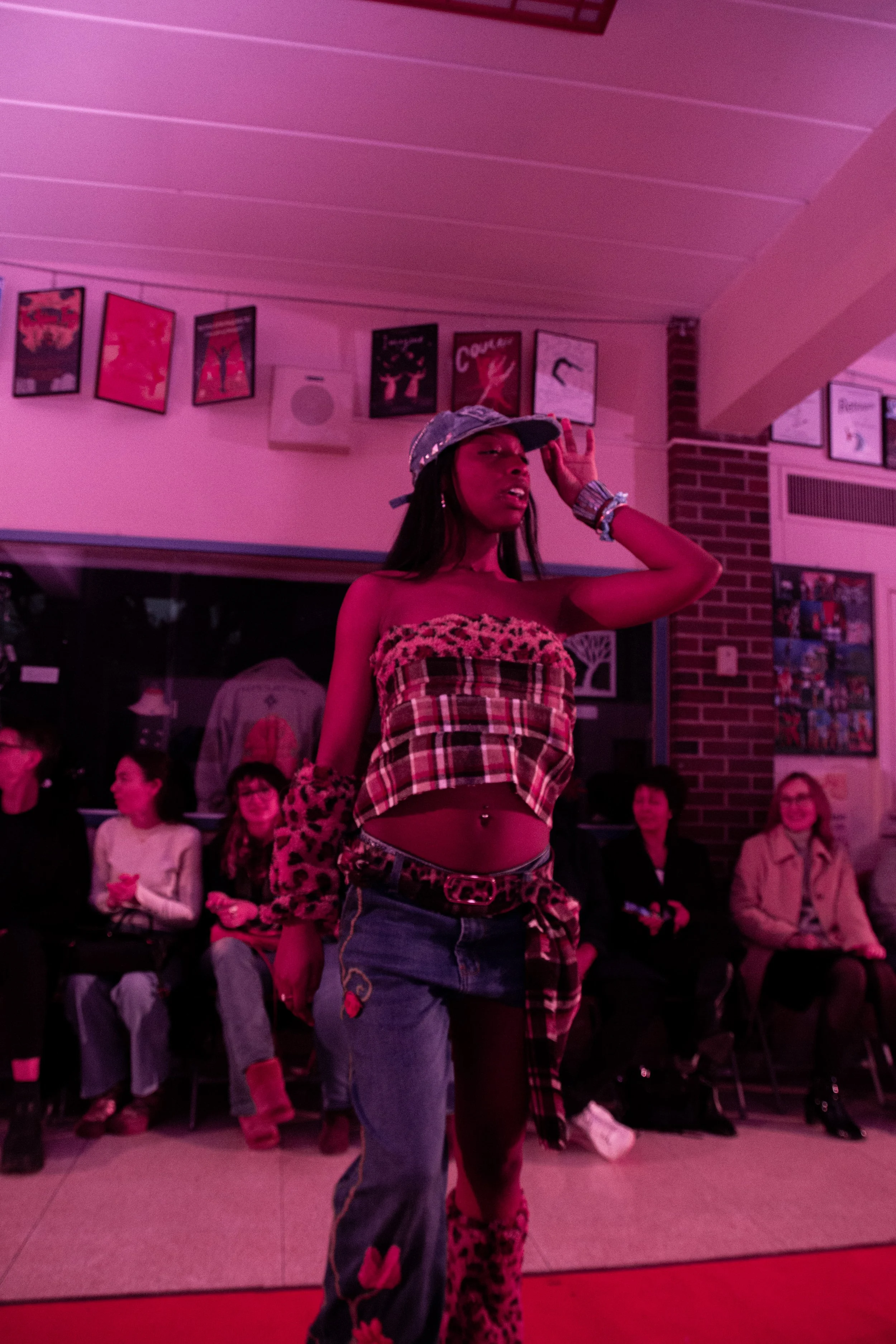 A woman standing with a hand on her head in front of an audience at an indoor event with pink lighting and framed pictures on the wall.