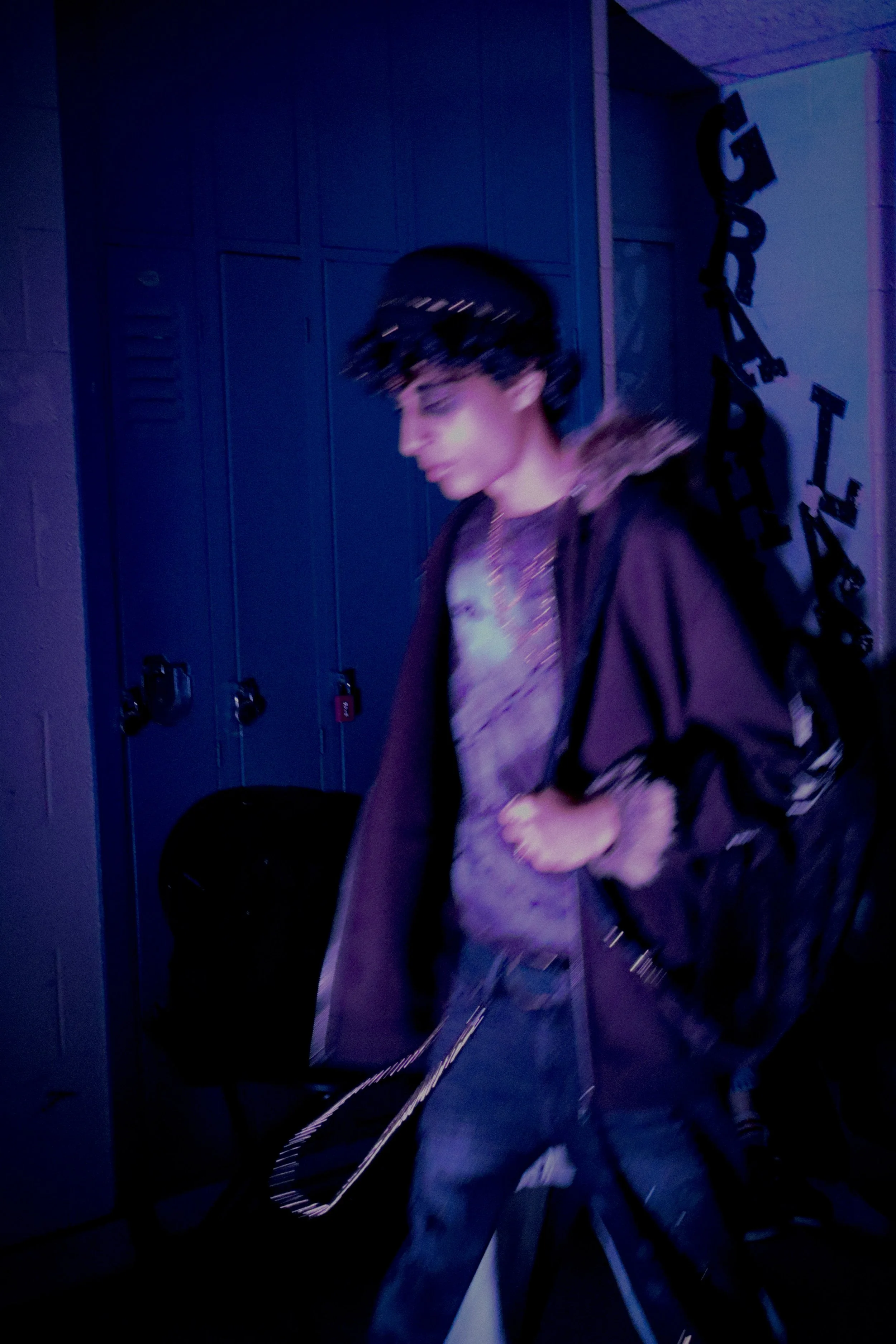 A person with dark hair and light skin walking in a dimly lit room, possibly a school or gym locker room, with blue lockers and a sign that reads 'GARE' in the background.