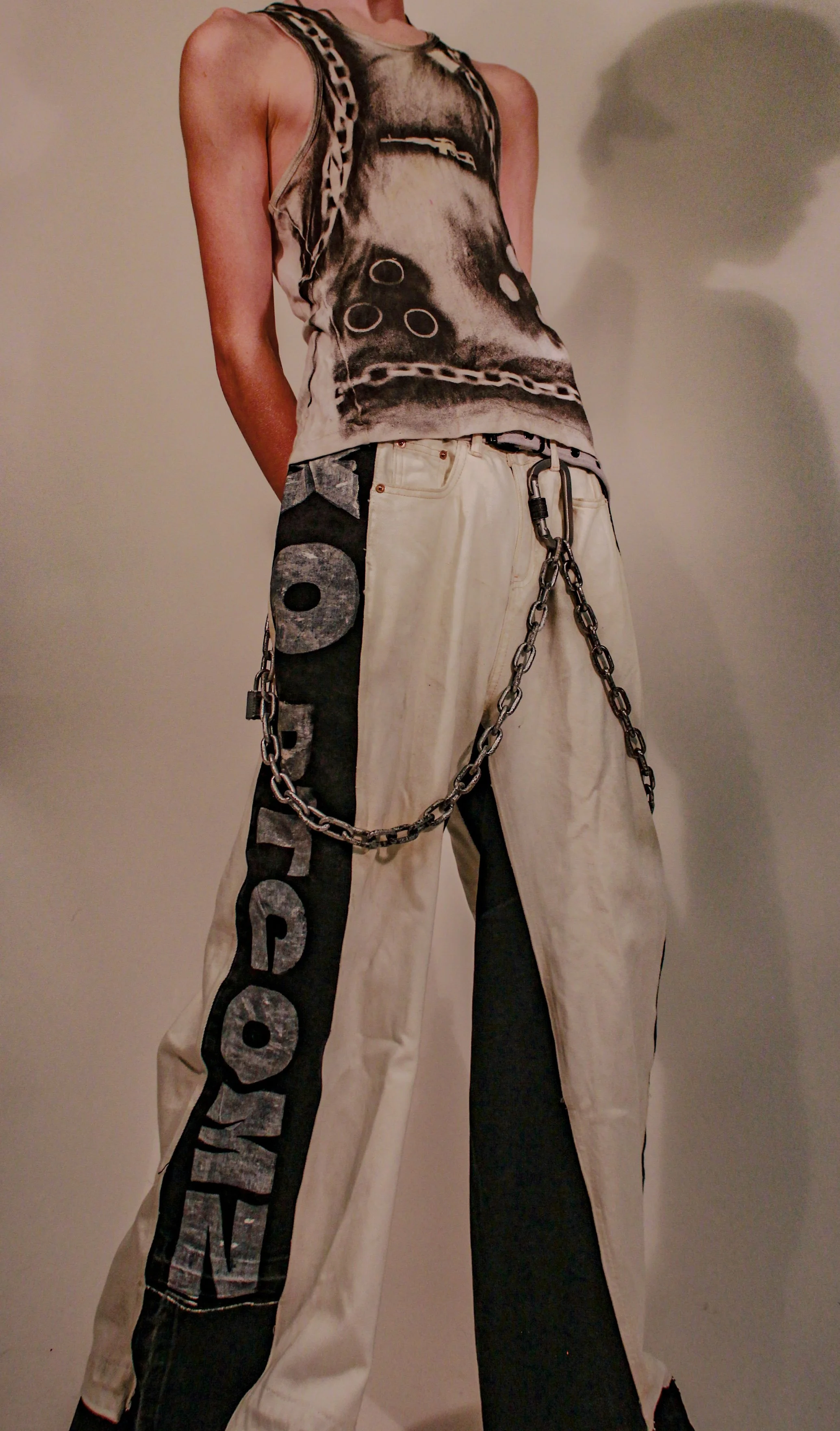 Person wearing wide-legged beige pants with black side stripe, a chain hanging from the belt loop, and a sleeveless graphic tank top featuring a dog face with circular designs, standing against a plain wall.