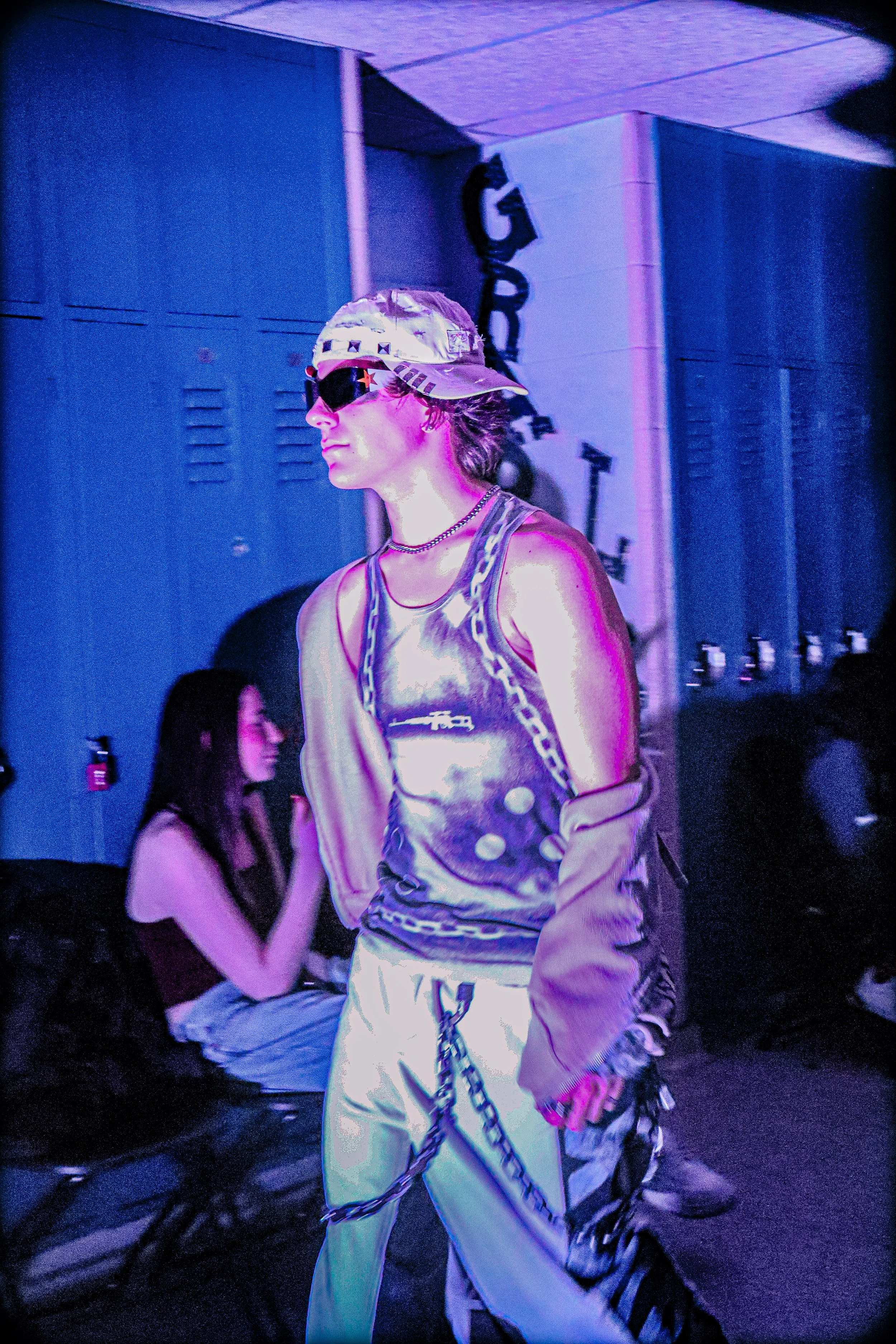 Person walking in a dimly lit room illuminated by purple and blue lighting, wearing sunglasses, a shiny metallic vest, a beige cap, and white pants with black and white striped accents.