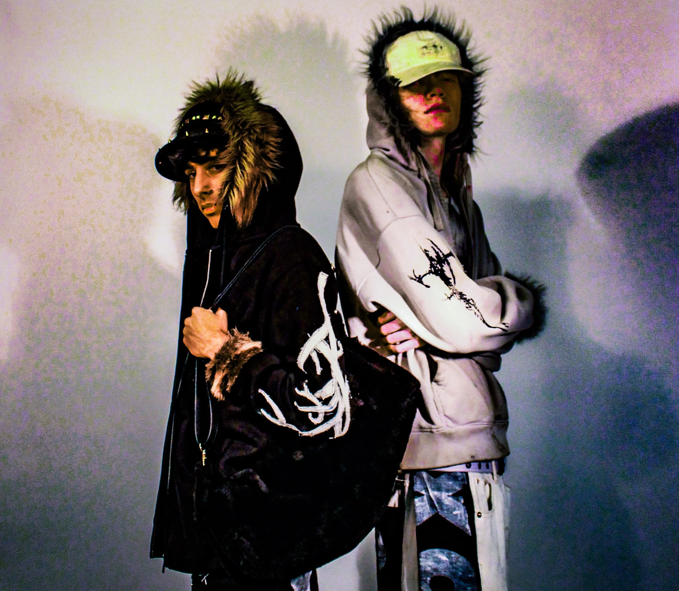 Two young men standing side by side against a plain wall, wearing hooded jackets with fur-lined hoods and baseball caps, looking serious and stylish.