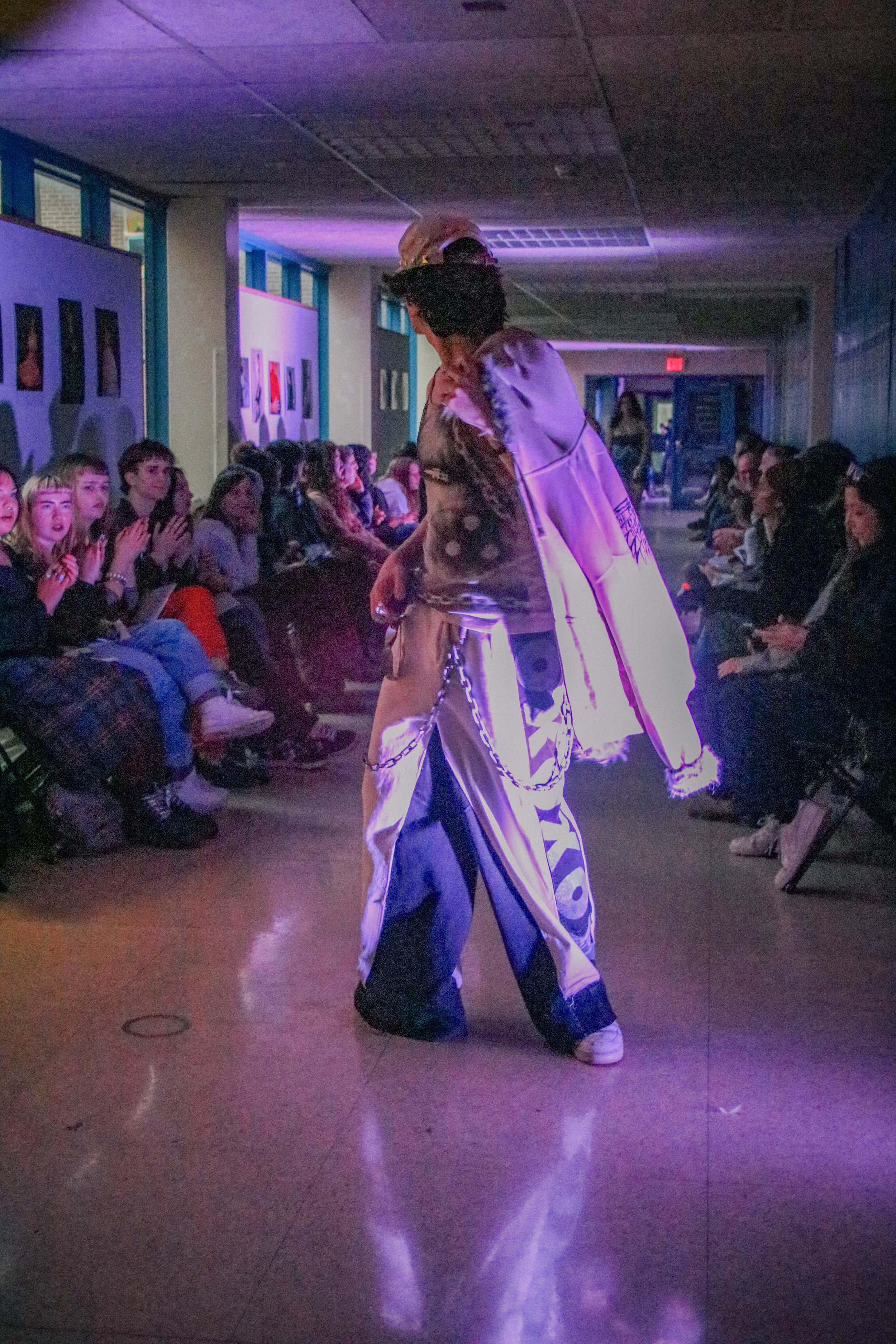 A person wearing a helmet and oversized clothing dances on a reflective floor in front of an audience watching in a dimly lit room decorated with artwork on the walls.
