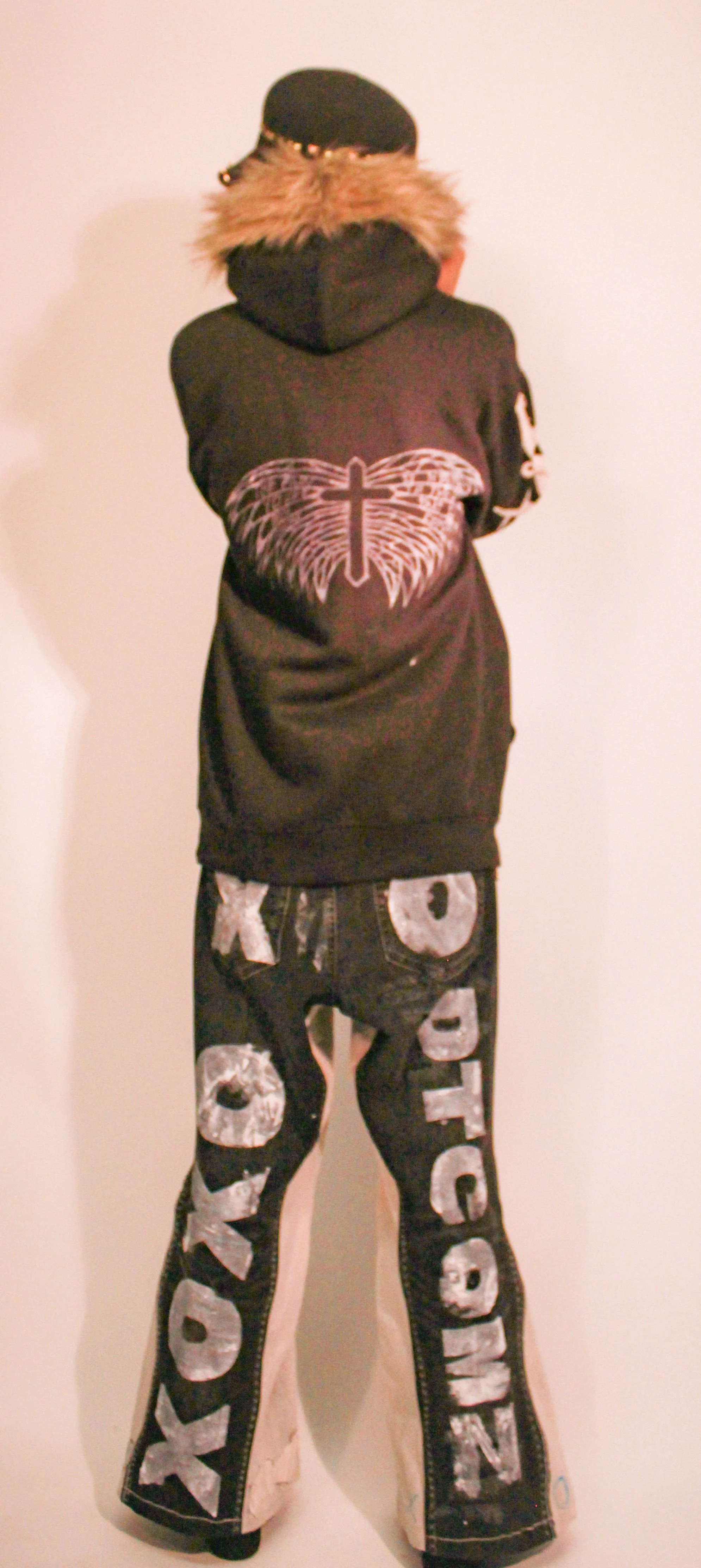 Back of a person wearing a black hoodie with a cross and angel wings design on the back, black pants with large white letters spelling 'ROCK' down the legs, and a black cap with a fur-lined hood.