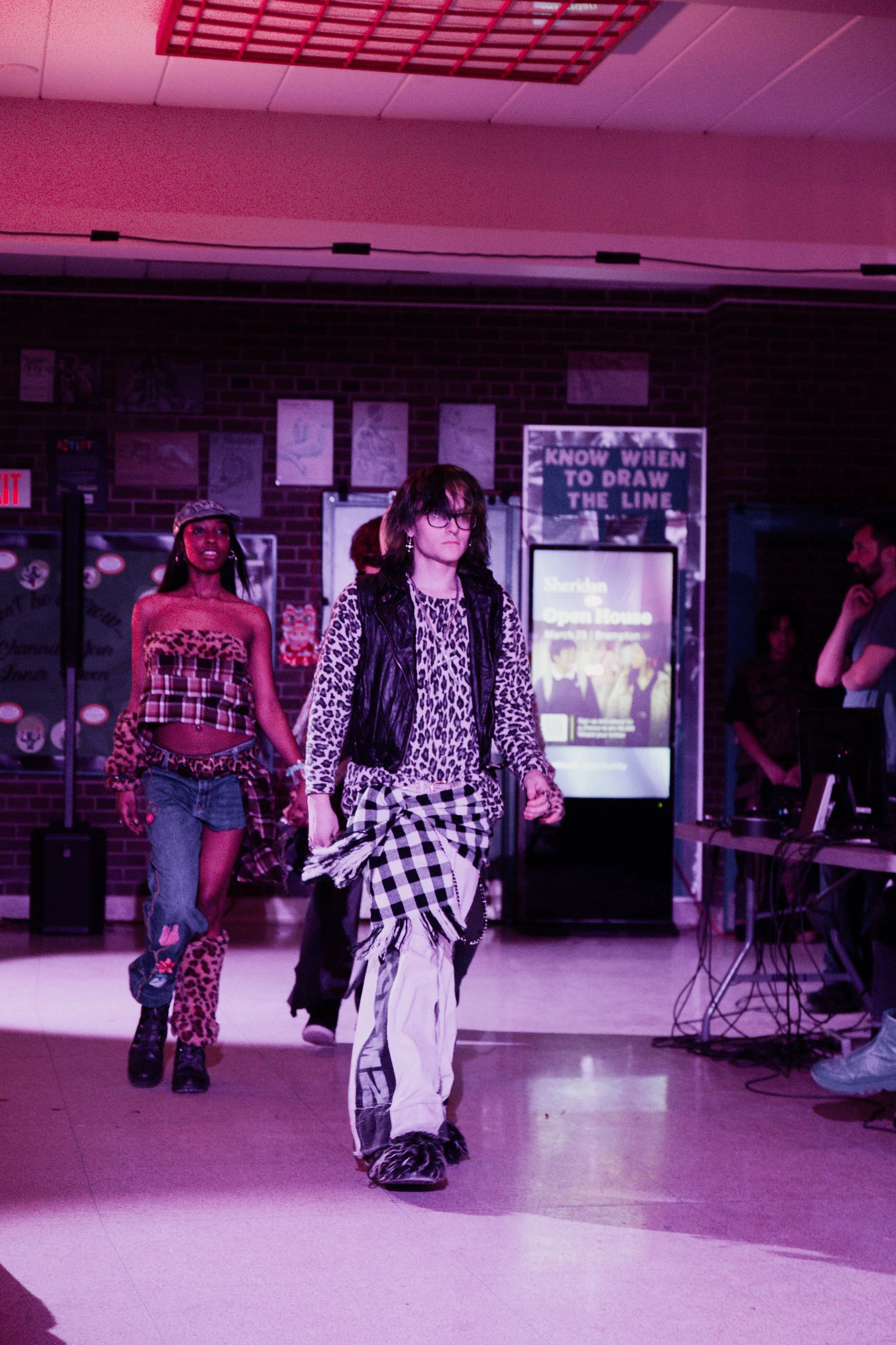 Three women walking on a runway with brick walls and posters in the background, pink lighting, and electronic equipment on the side.