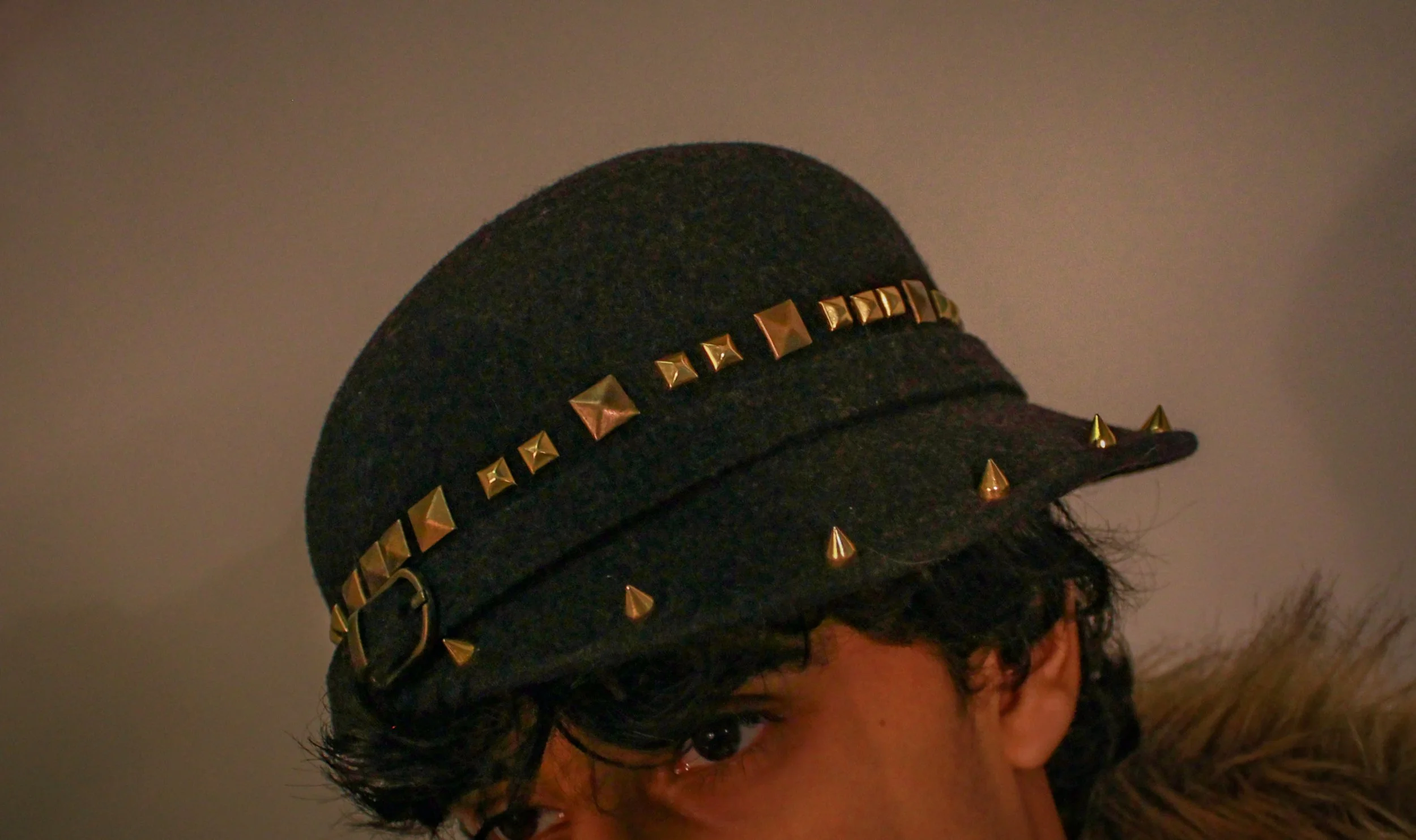 Close-up of a person wearing a black cap decorated with gold pyramid-shaped studs, with dark hair partially visible and a gray wall in the background.