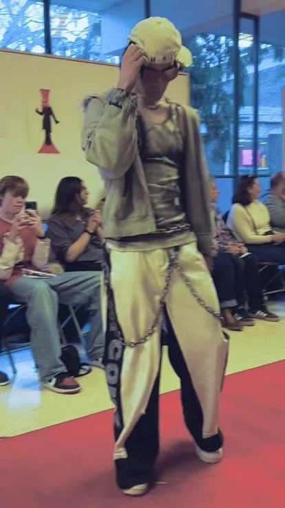 A person dressed in a casual outfit with a chain hanging from their pants, holding a yellow object or hat to their head, in a room with seated audience members and colorful decor.